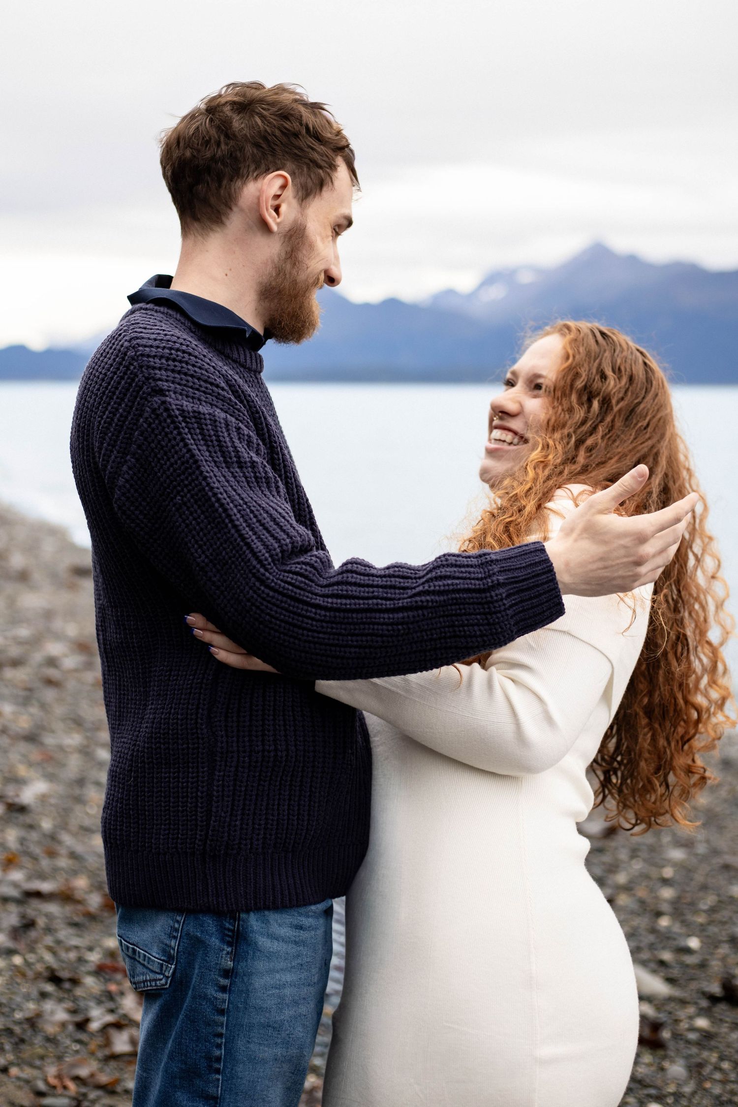 A couple embraces joyfully on a rocky beach with mountains in the background.