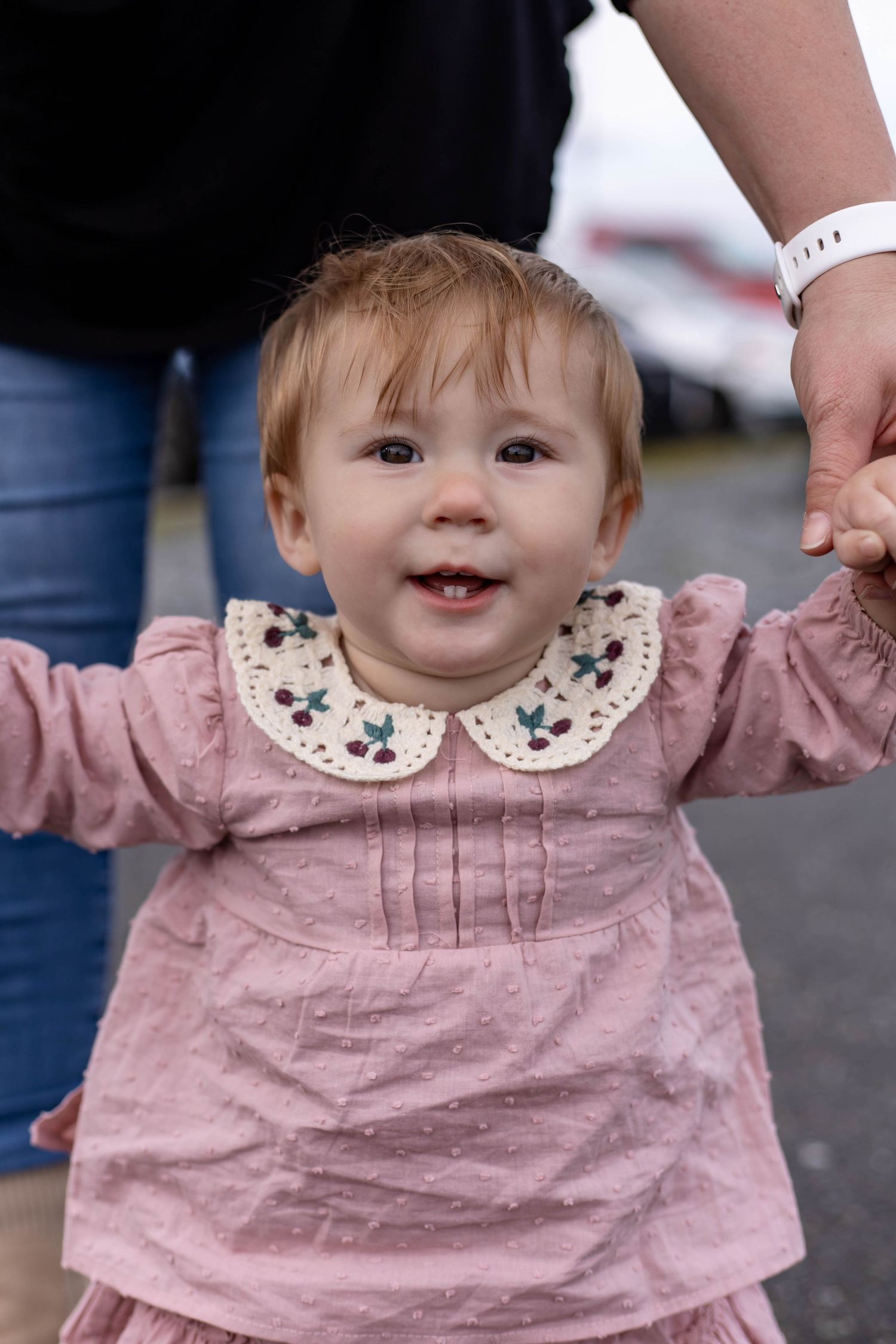 Baby in pink dress with floral collar smiles while being held up by supporting hands.
