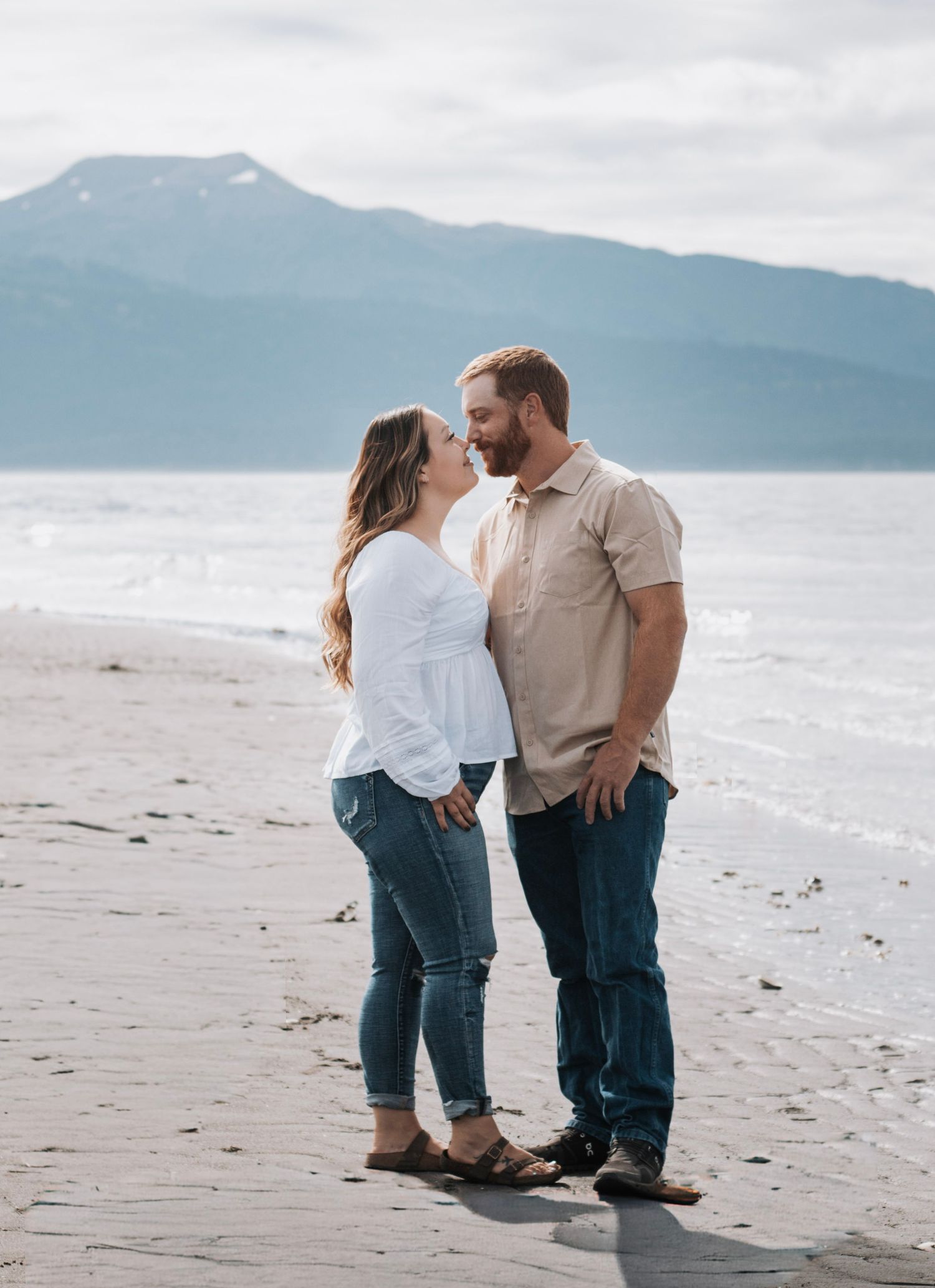 Couple in casual attire sharing tender moment on beach with mountains and ocean backdrop.