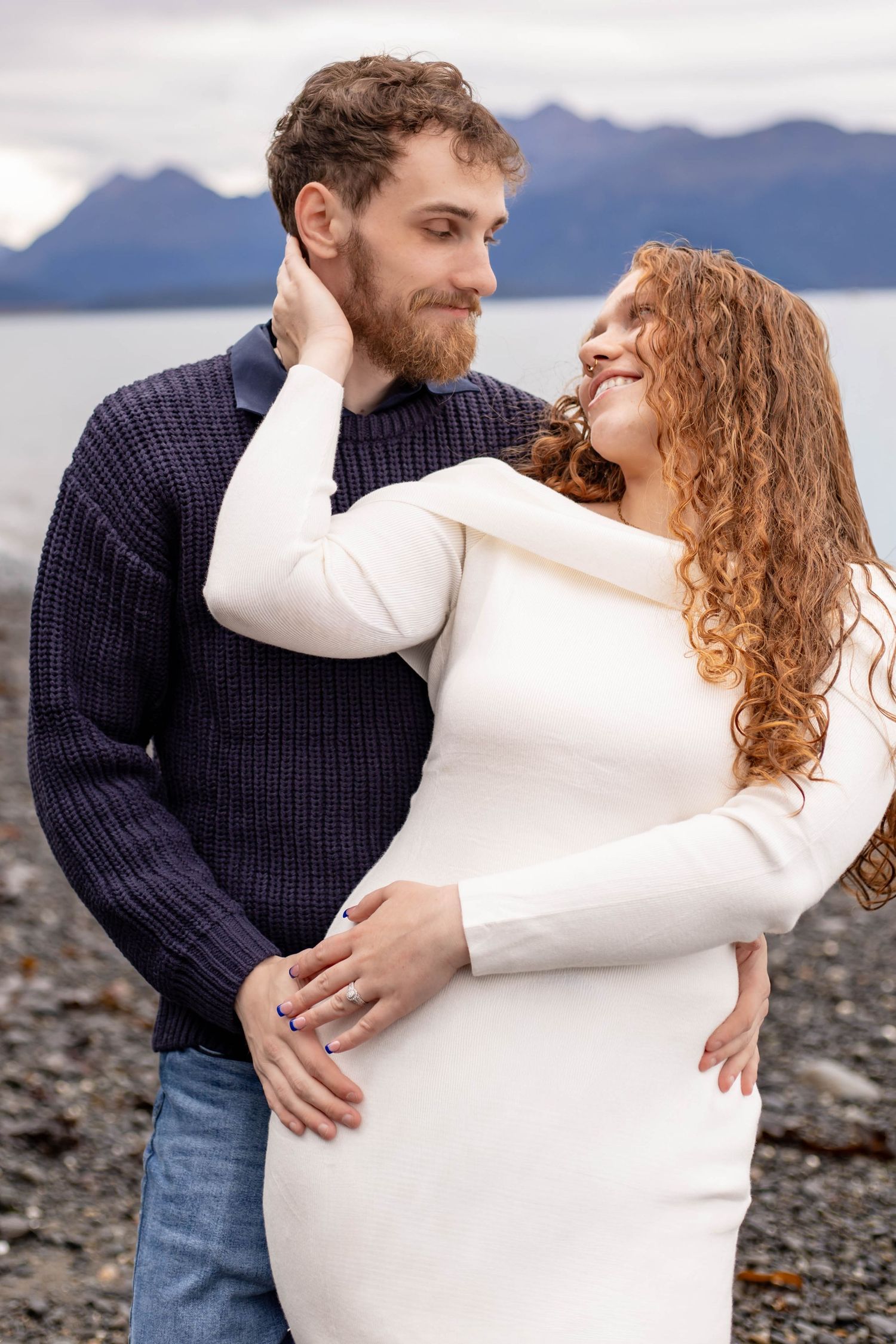 A romantic couple embraces on a rocky beach with mountains and water in the background.