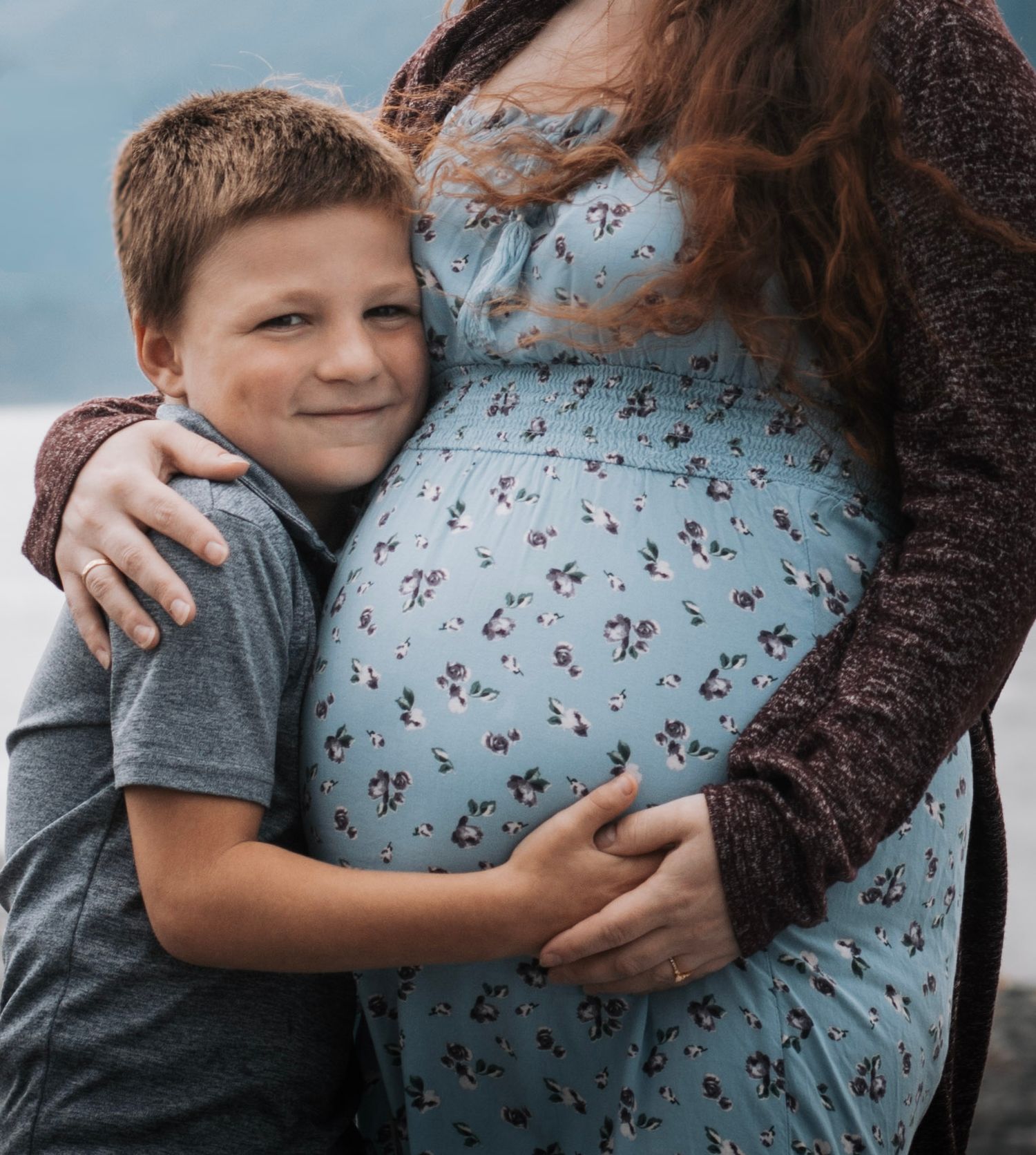A young boy smiles while hugging a pregnant woman outdoors near water.