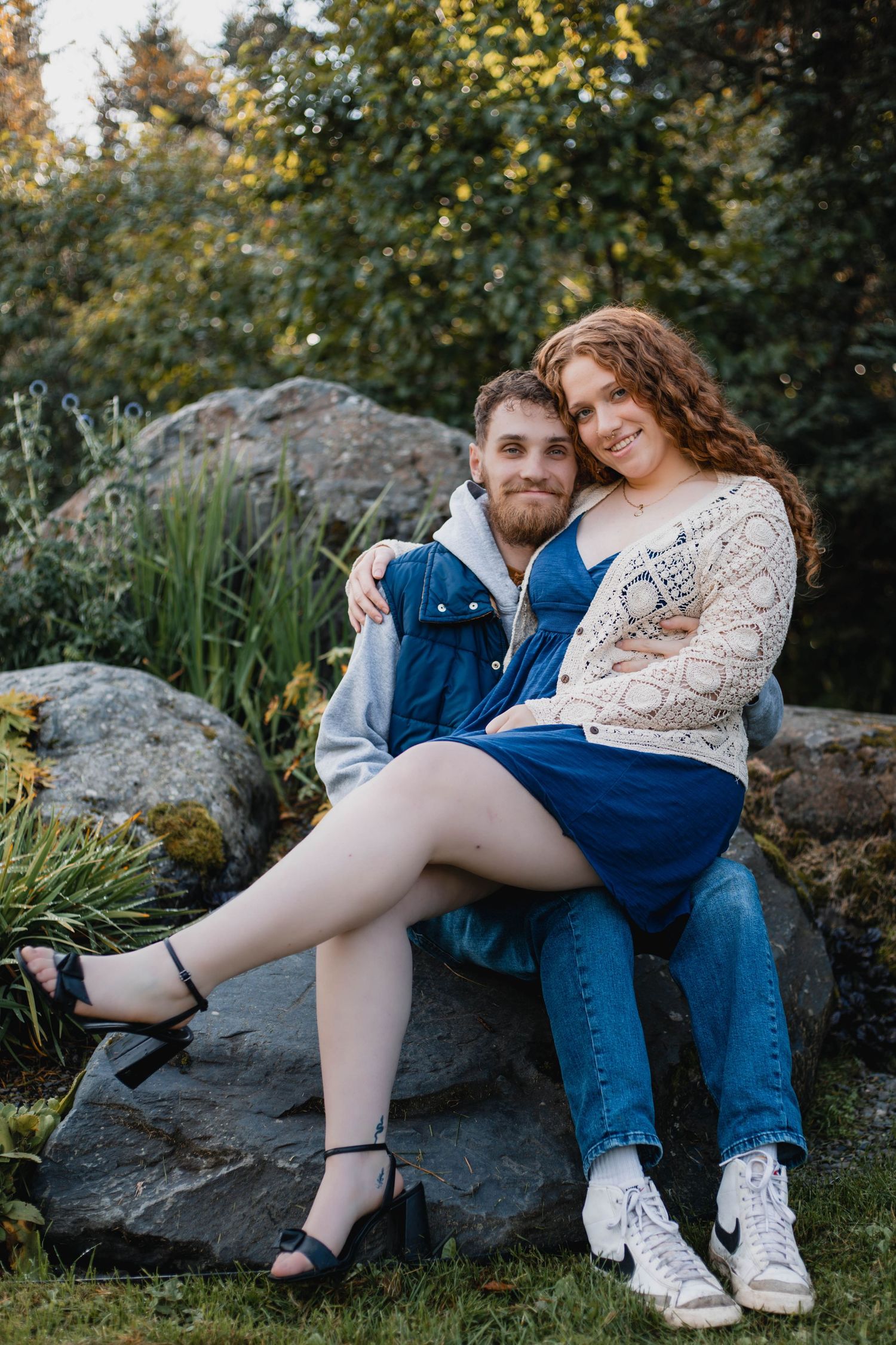 A romantic couple sits together on large rocks in a garden setting during a photoshoot.