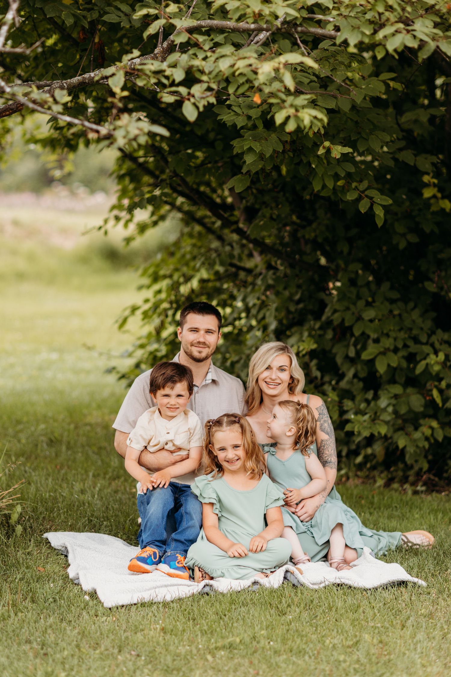 A family sits together on a white blanket under leafy green trees on a summer day.
