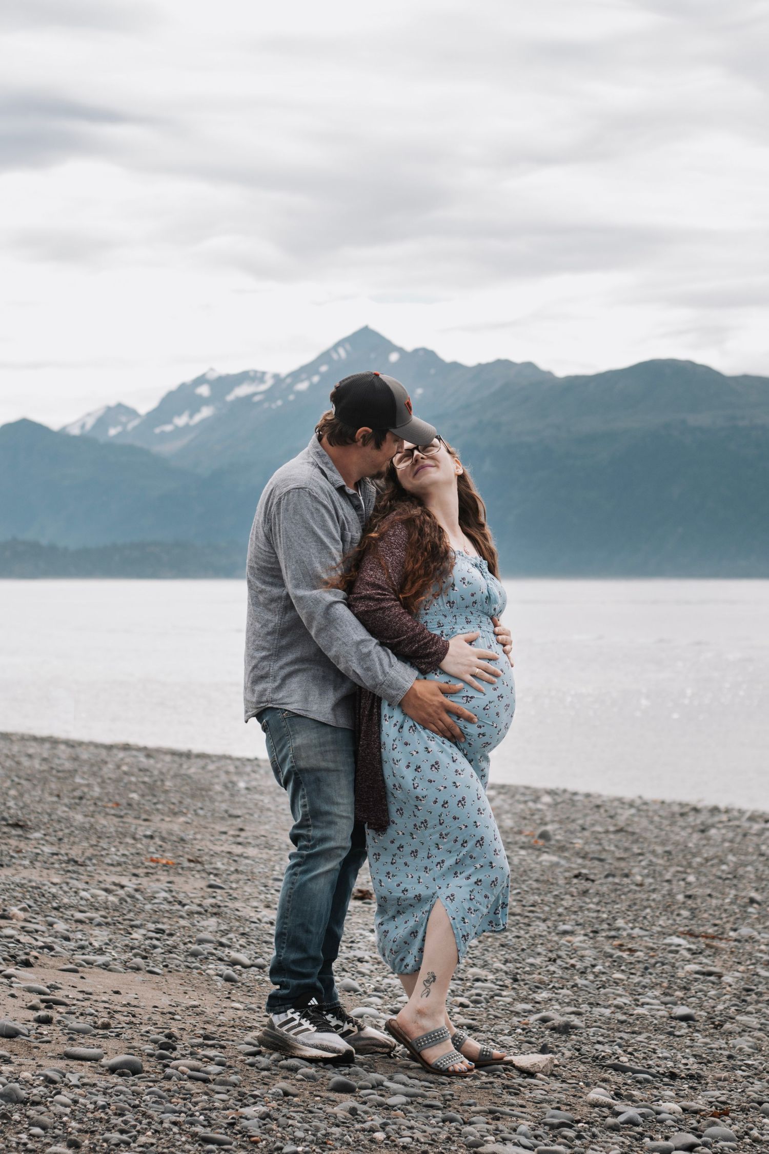 A couple shares romantic moments on a rocky beach with mountains and water in the background.