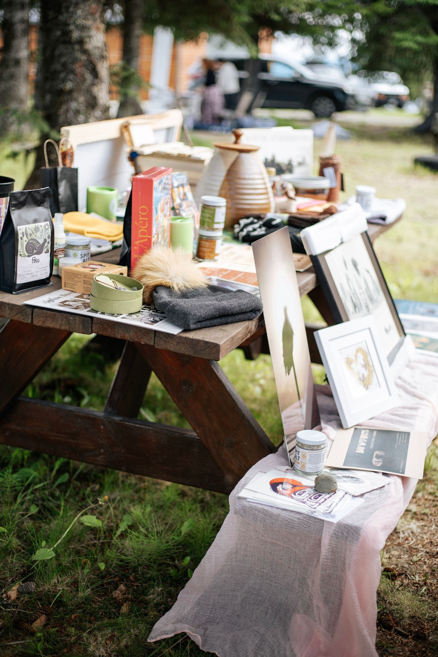 Outdoor market picnic table displaying art prints, ceramics, candles, and handmade goods at a craft fair.