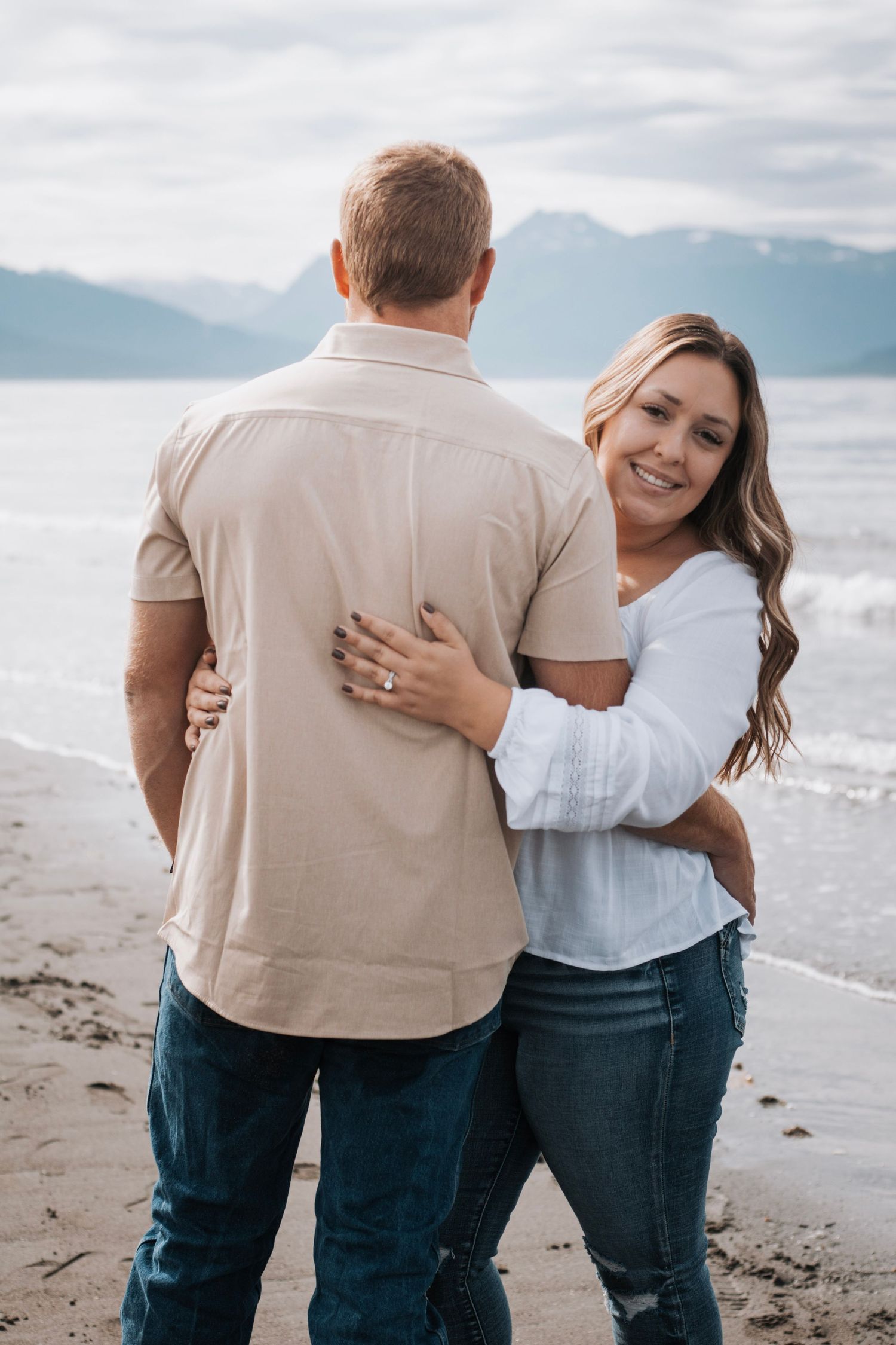 Couple embracing on beach wearing white and beige with mountains in background.