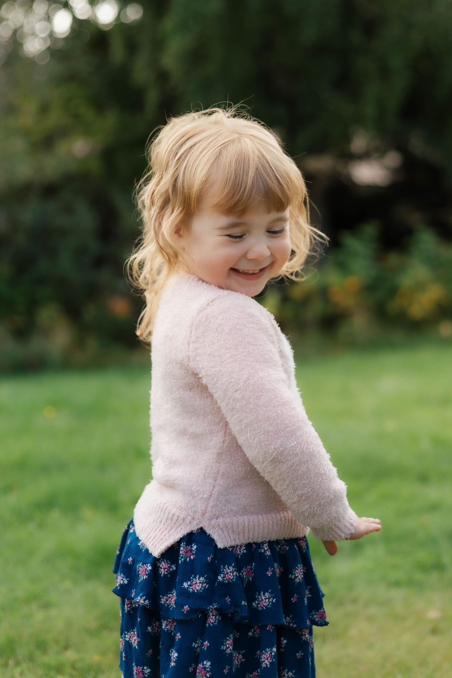 A child in a pink sweater and floral blue dress spins joyfully on a grassy lawn.