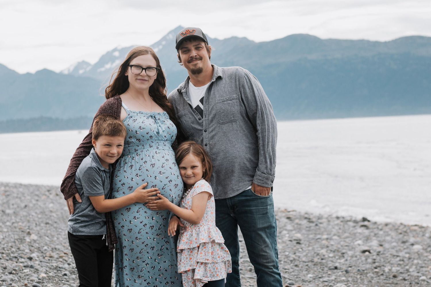 Pregnant family of four posing together on a rocky beach with mountain and water views.