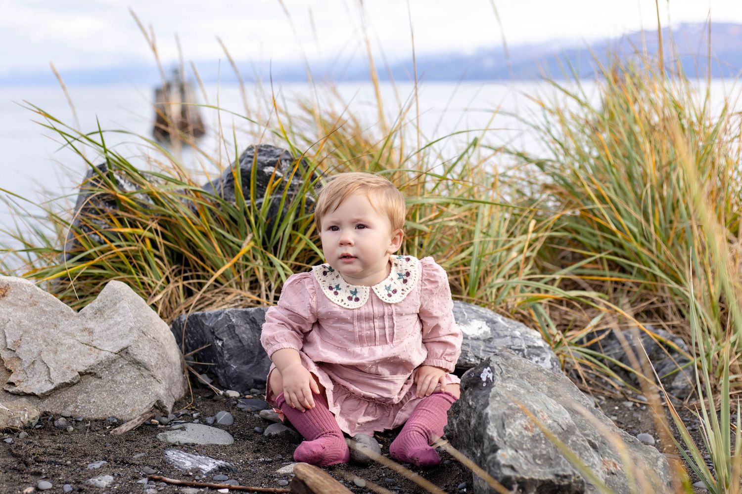 Two young children explore a rocky beach area with tall grass on a cloudy day.