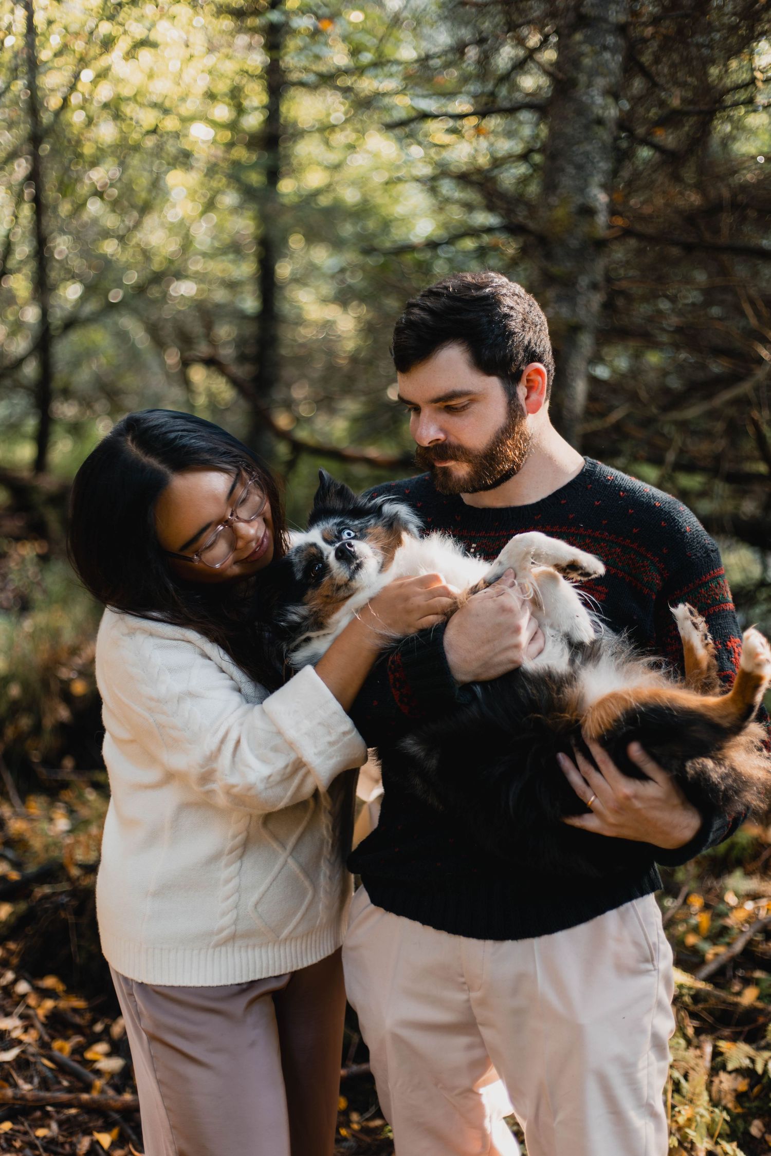 A couple in coordinating white and black outfits plays with their energetic dog in a forest setting during autumn.