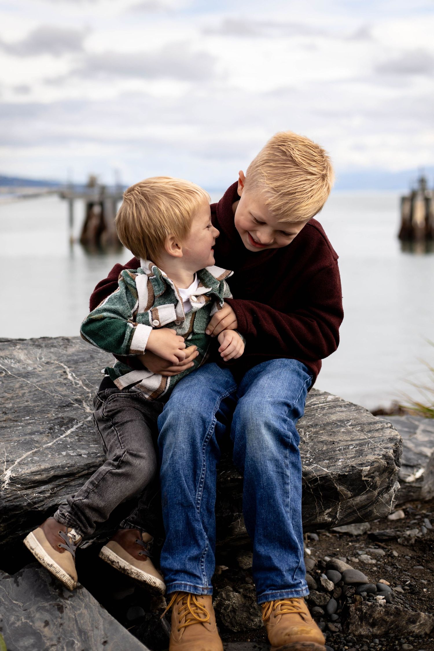 Two children share a playful moment while sitting on rocky shoreline near water.