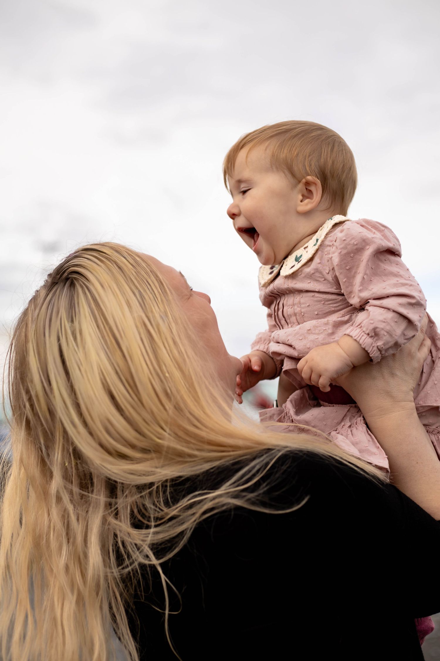 Blonde mother lifting laughing baby girl in pink dress outdoors against cloudy sky.