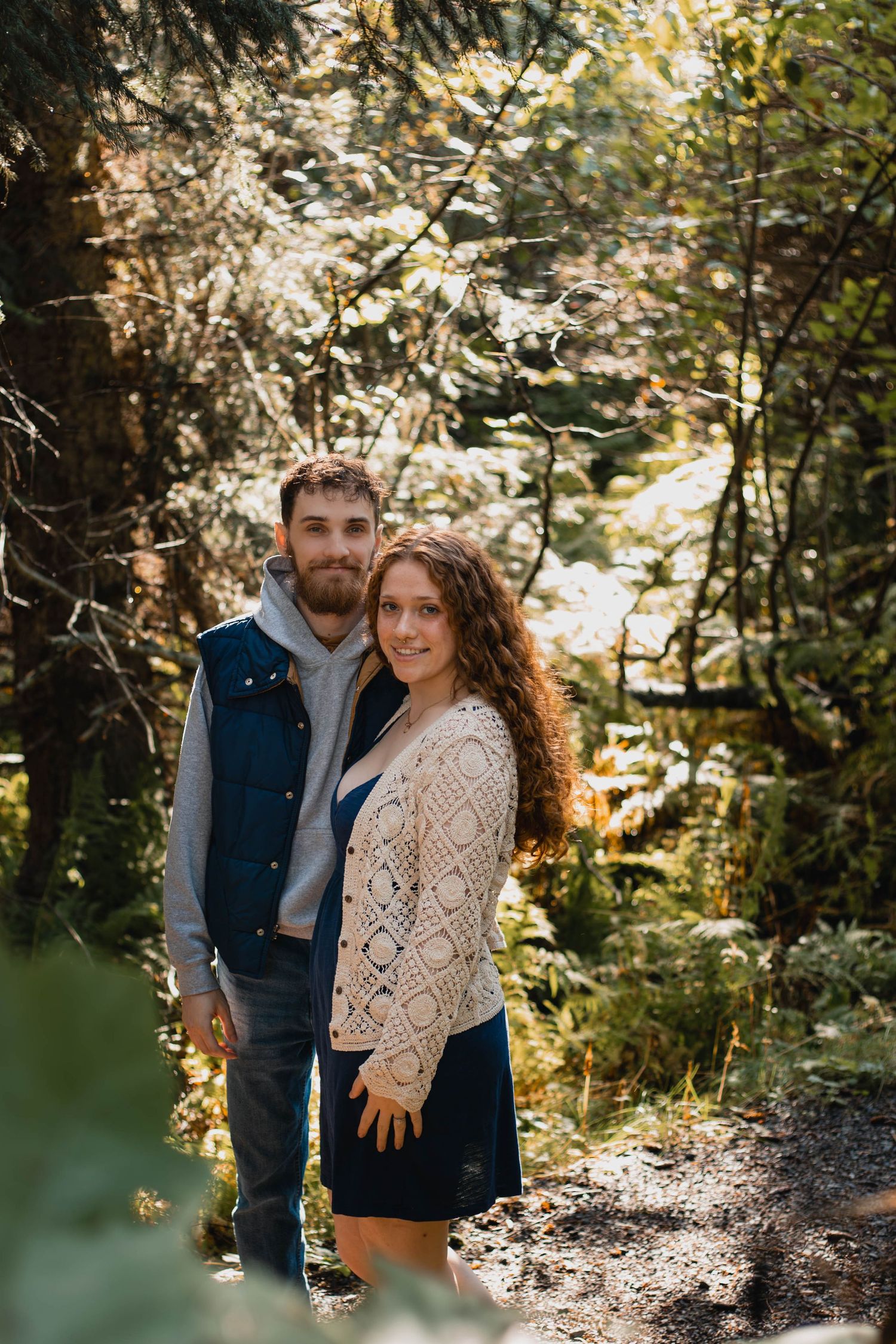 A couple embraces in a sunlit forest setting wearing casual autumn attire.