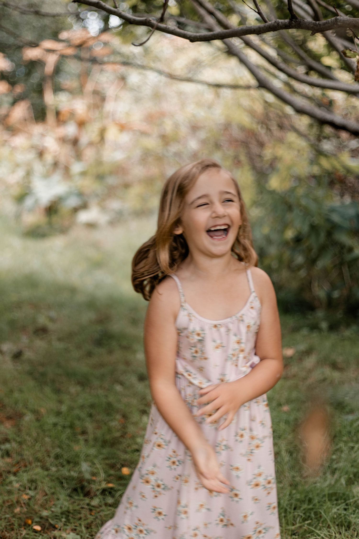 A child wearing a floral sundress laughs joyfully in a natural outdoor setting under trees.