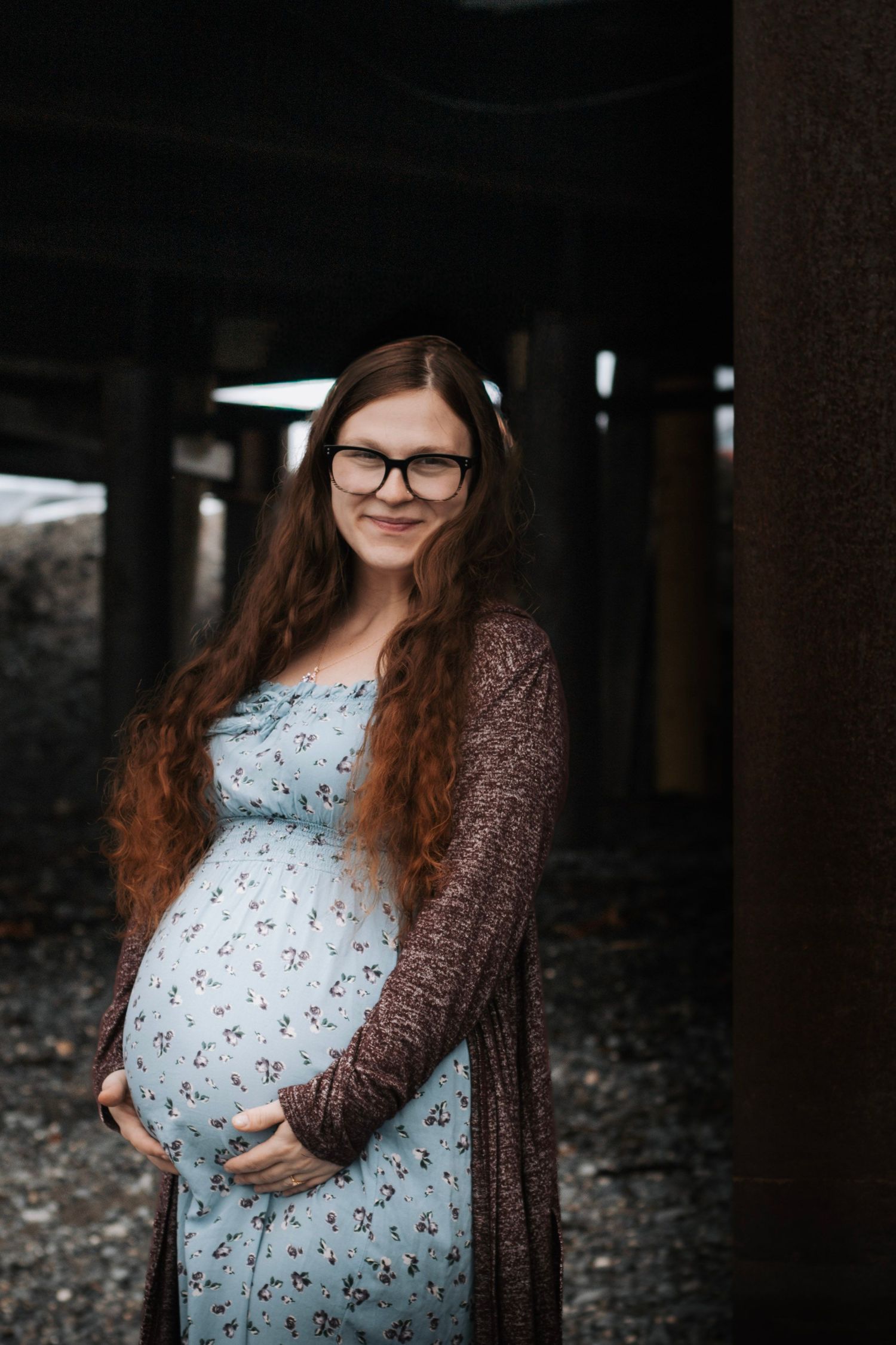 Smiling pregnant woman with long red hair and glasses in floral dress holding baby bump under pier.