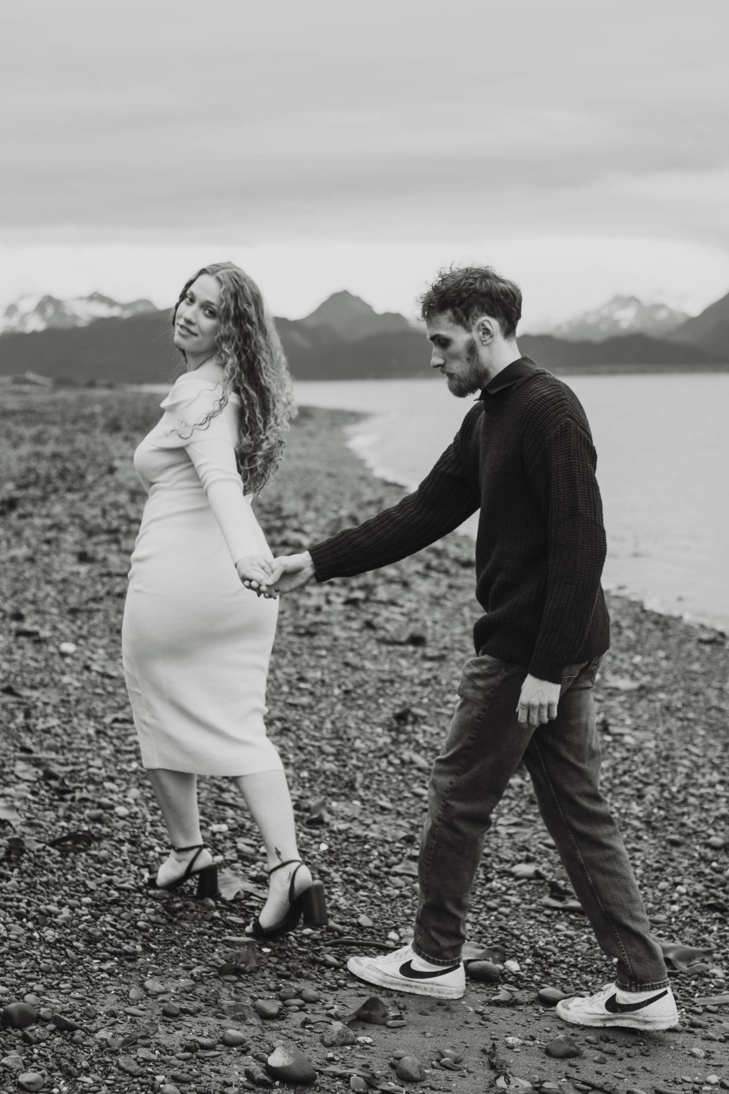 Playful couple in casual wear dancing on a rocky shoreline with mountains visible in the distance.