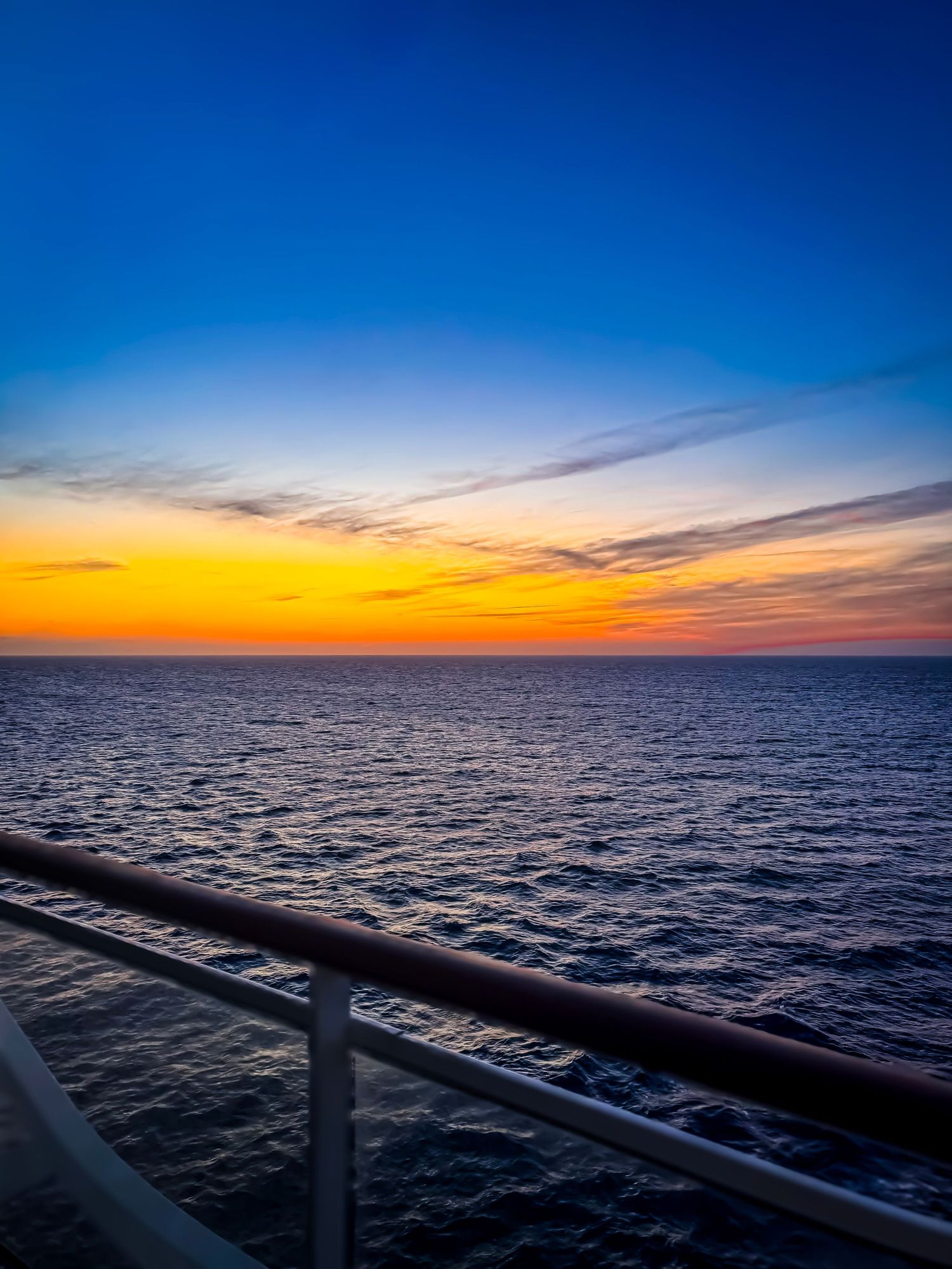 The dark ocean waters stretch to the horizon under a vibrant orange and blue sunset sky from a cruise ship deck railing view.