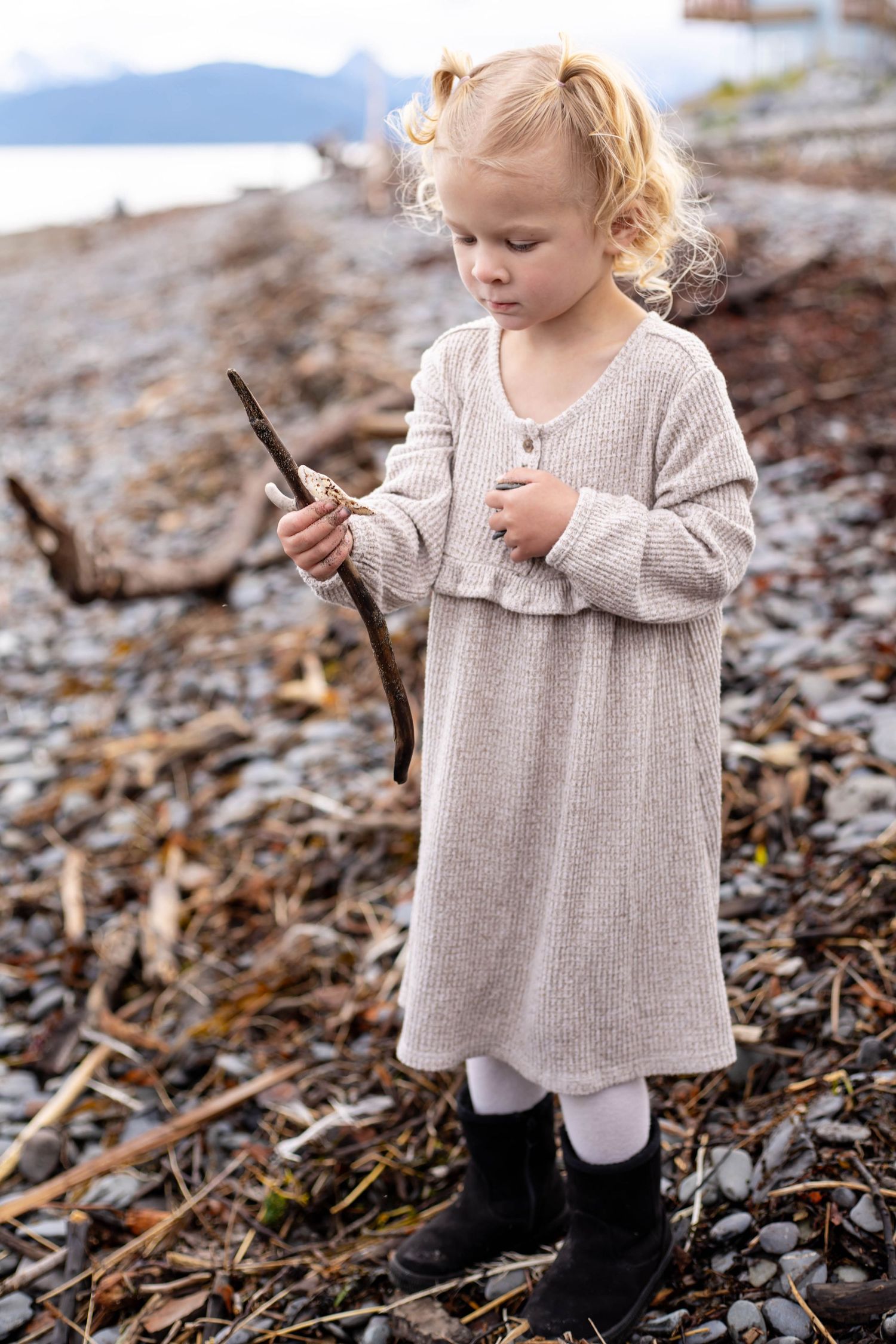 A small child in a beige sweater explores fallen leaves and driftwood on a rocky beach.