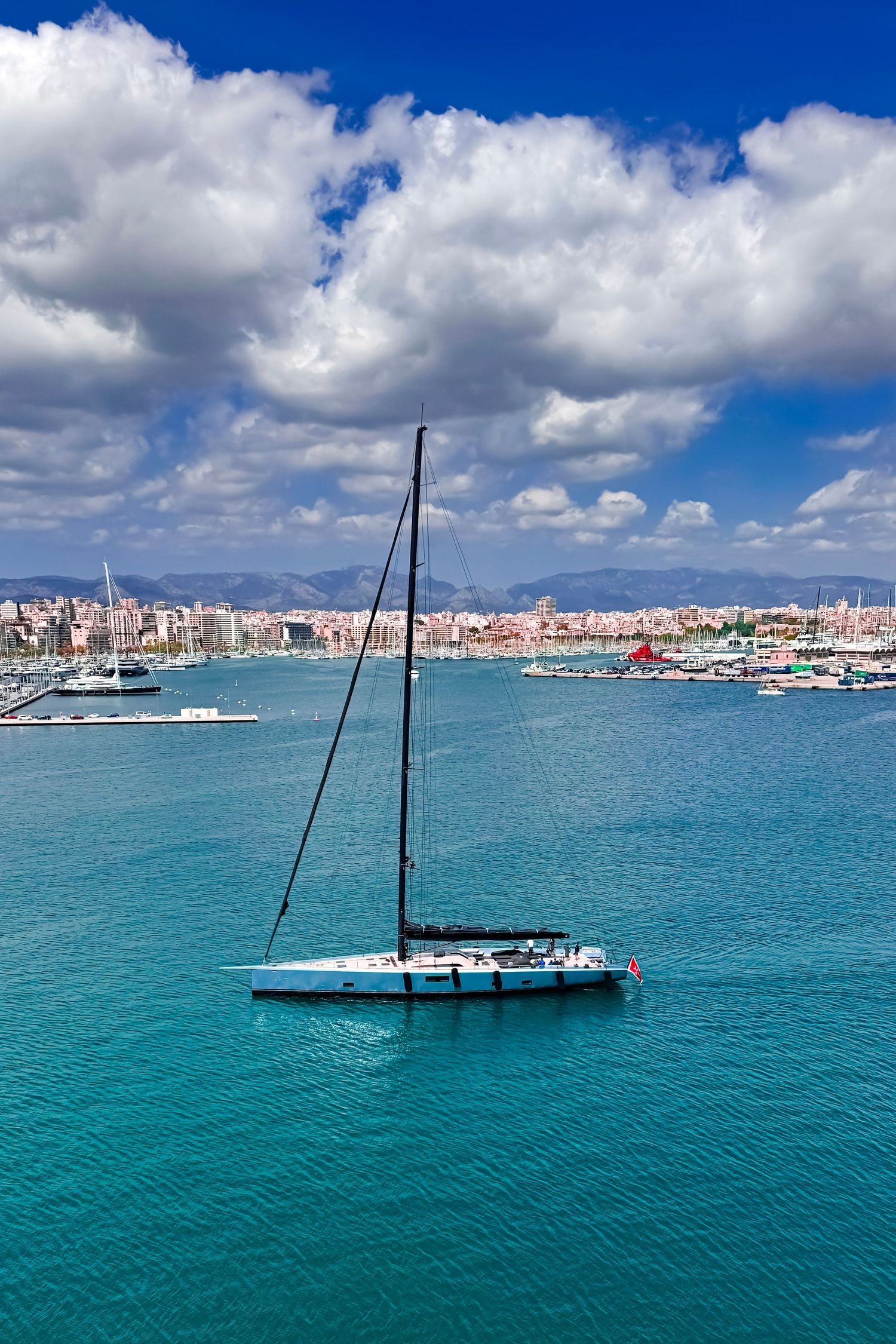 Sailboat sits partially submerged with mast protruding from turquoise Mediterranean waters near coastal town harbor.