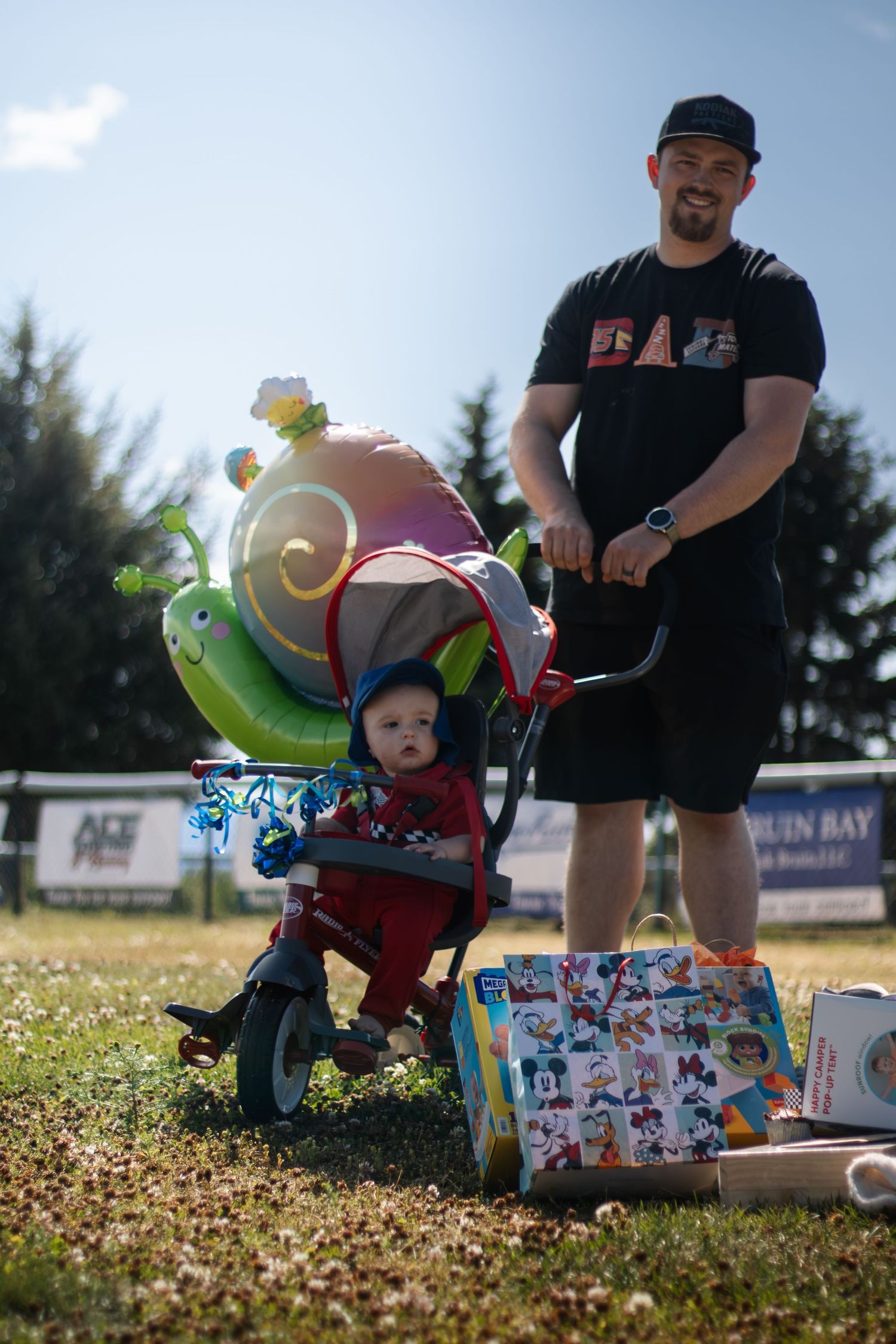 Man pushing toddler on tricycle with snail balloon outdoors, surrounded by wrapped birthday gifts on grass.
