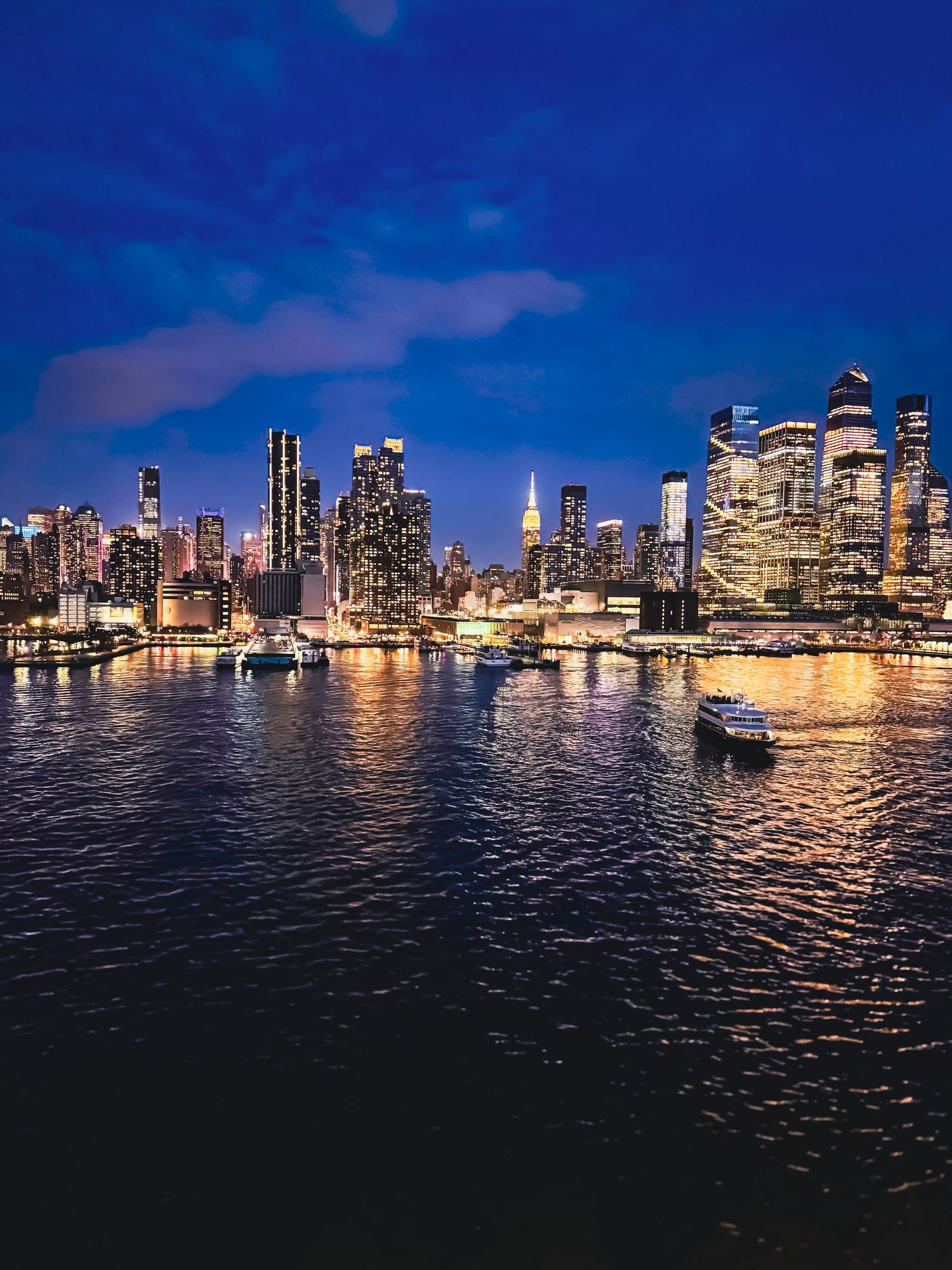 Manhattan skyline illuminated at dusk with glowing city lights reflecting on East River's dark waters.
