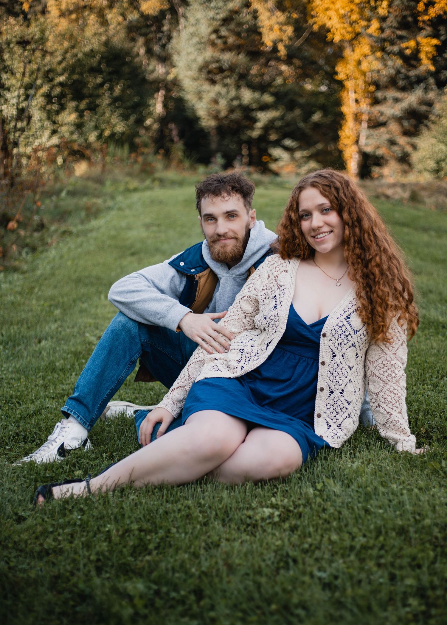 Casual outdoor portrait of couple sitting on grass with golden autumn foliage in background.