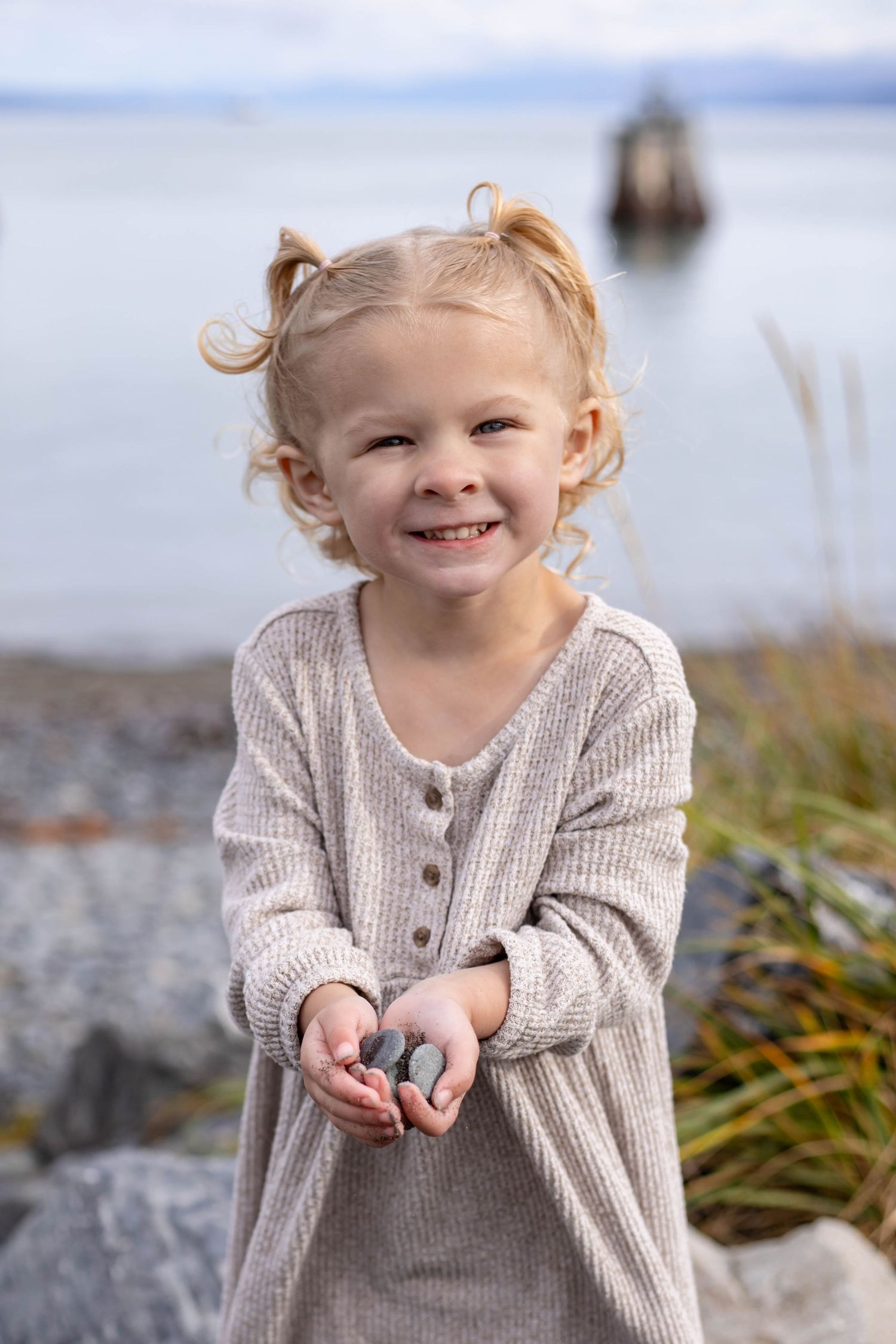 A young child in a beige sweater smiles while holding small objects at the beach.