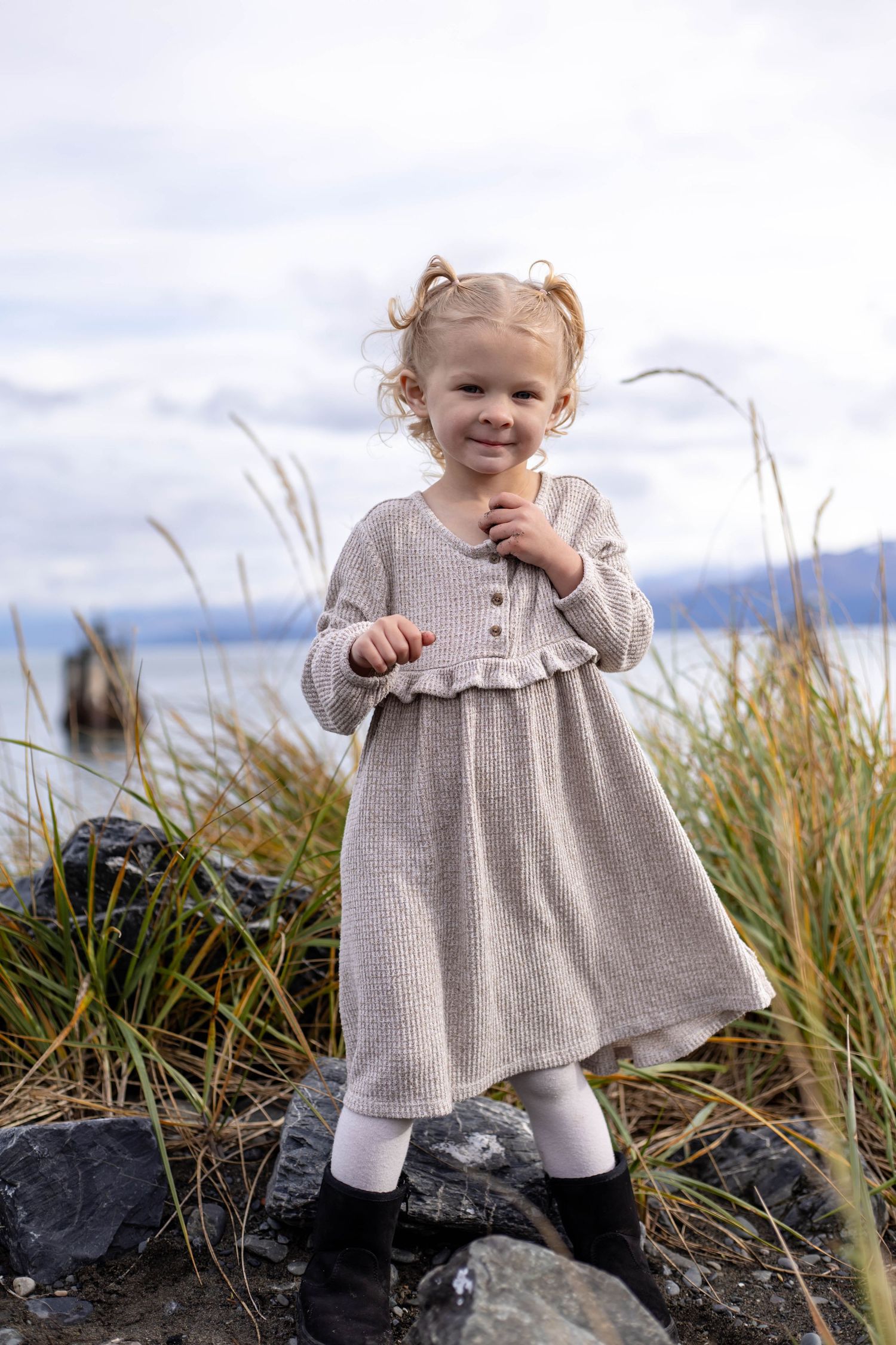 A child in a gray dress stands on coastal rocks surrounded by tall grass against a cloudy sky.