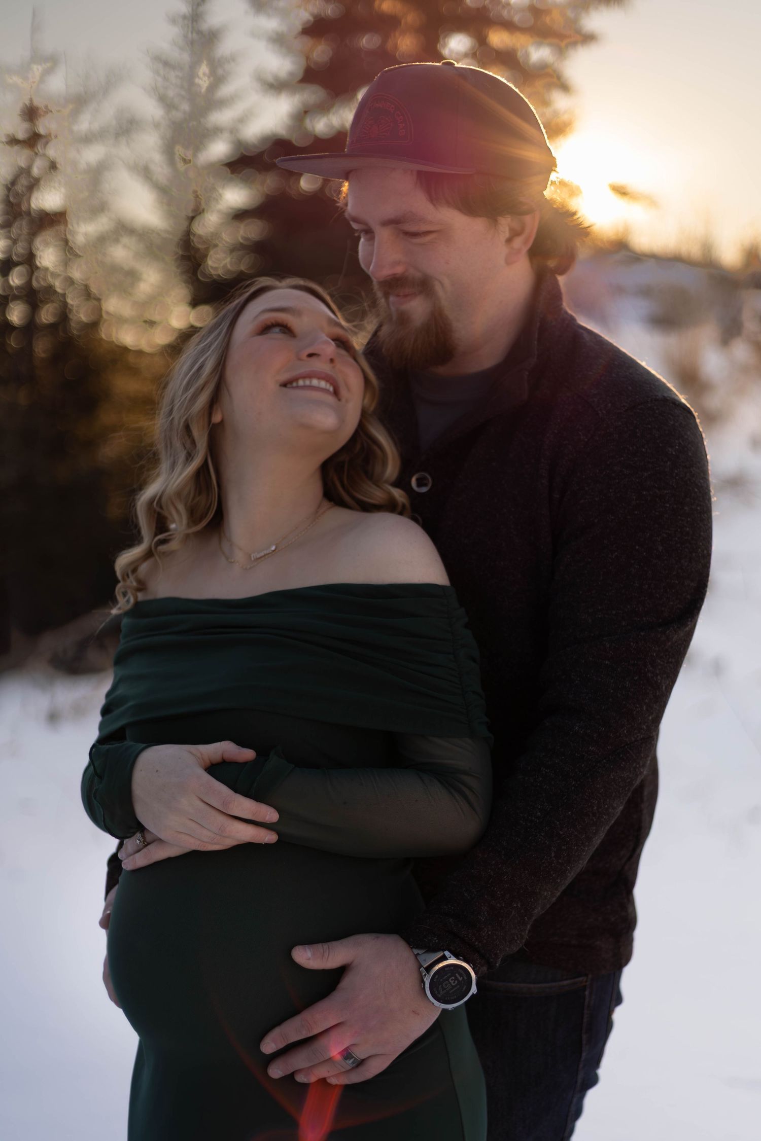 Couple embracing outdoors in snow at golden sunset, pregnant woman in green off-shoulder dress, man in hat and dark sweater.