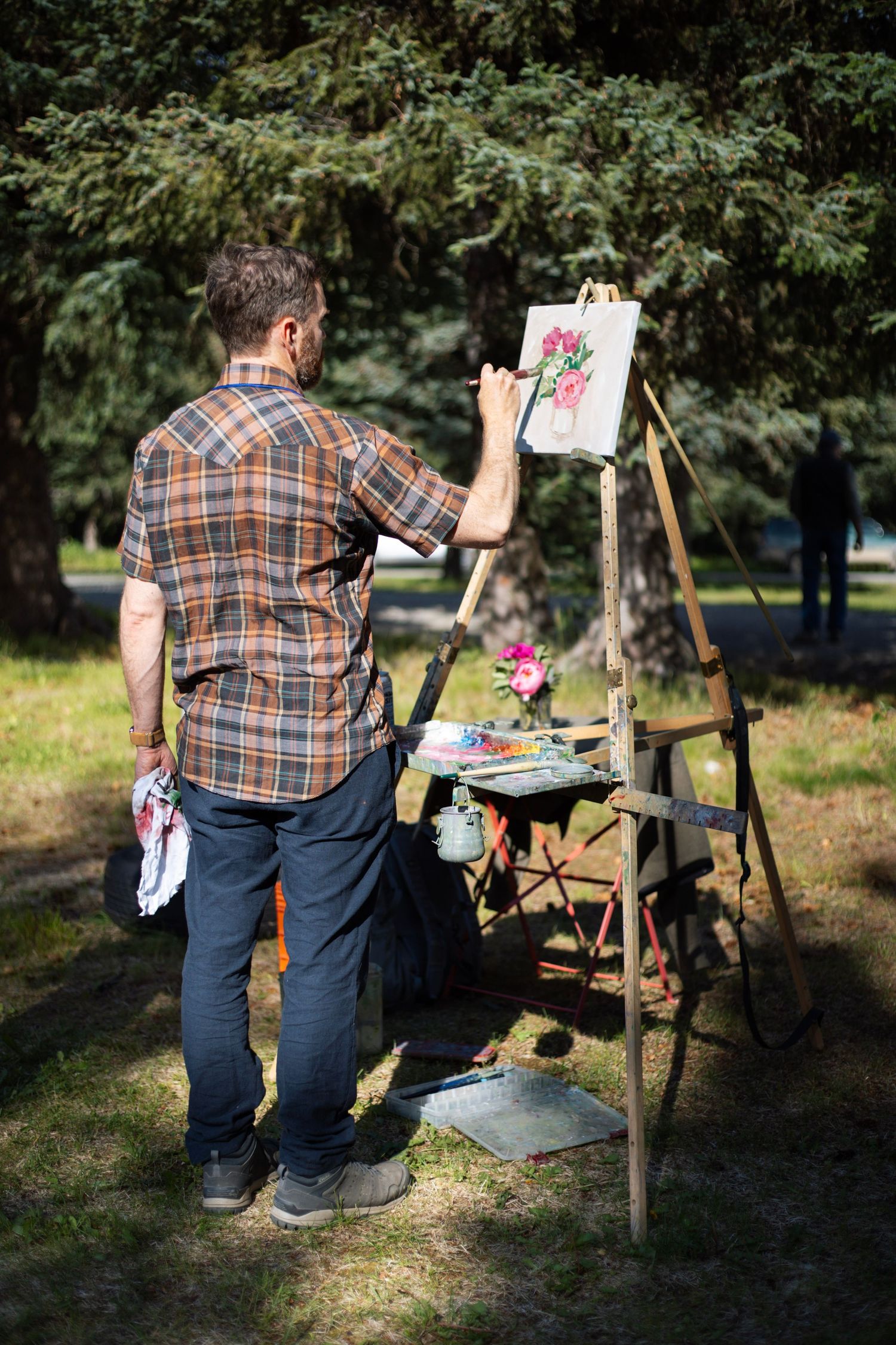 Man in plaid shirt painting floral artwork on easel outdoors in a sunny park surrounded by green trees.