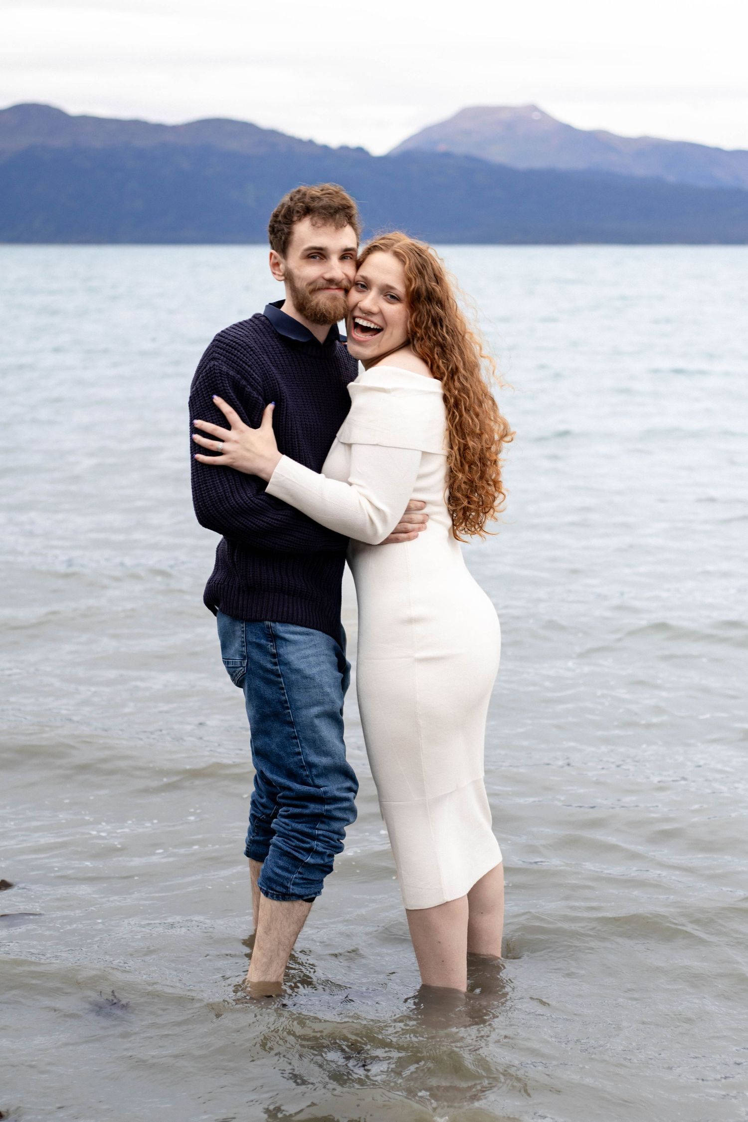 A couple embraces while standing in shallow water with mountains visible in the background at sunset.