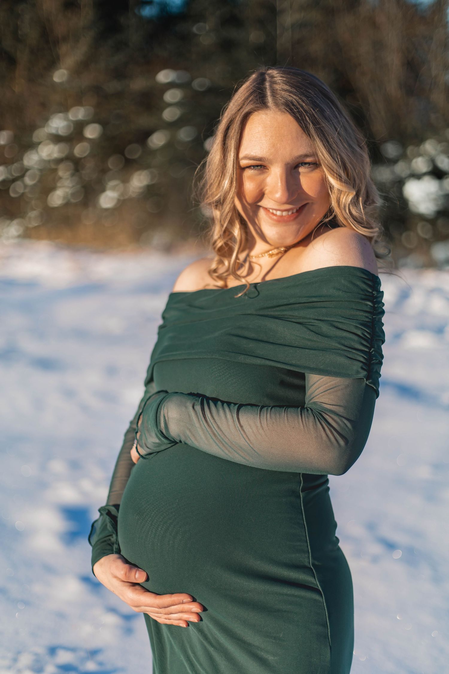 Smiling pregnant woman in green off-shoulder dress cradling baby bump outdoors in snowy winter setting.