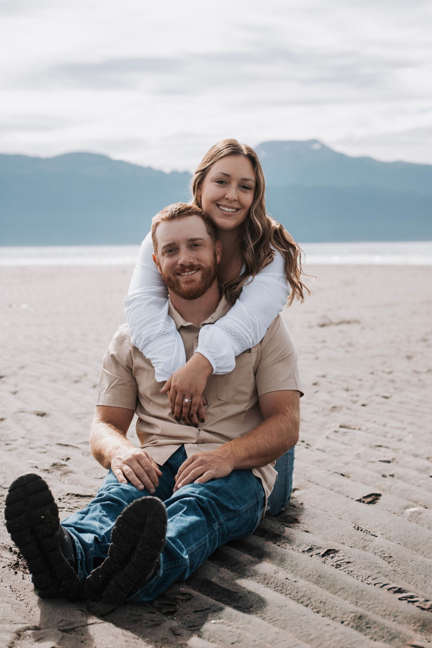 Couple relaxing together on sandy beach with mountains and ocean in background.