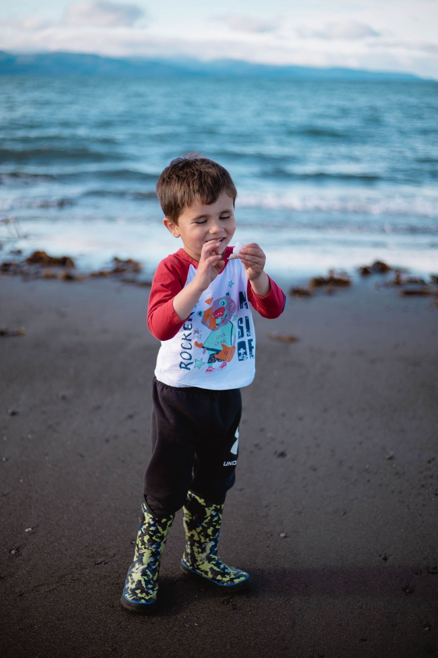 A young child in rain boots investigates items found on a misty beach at the ocean's edge.