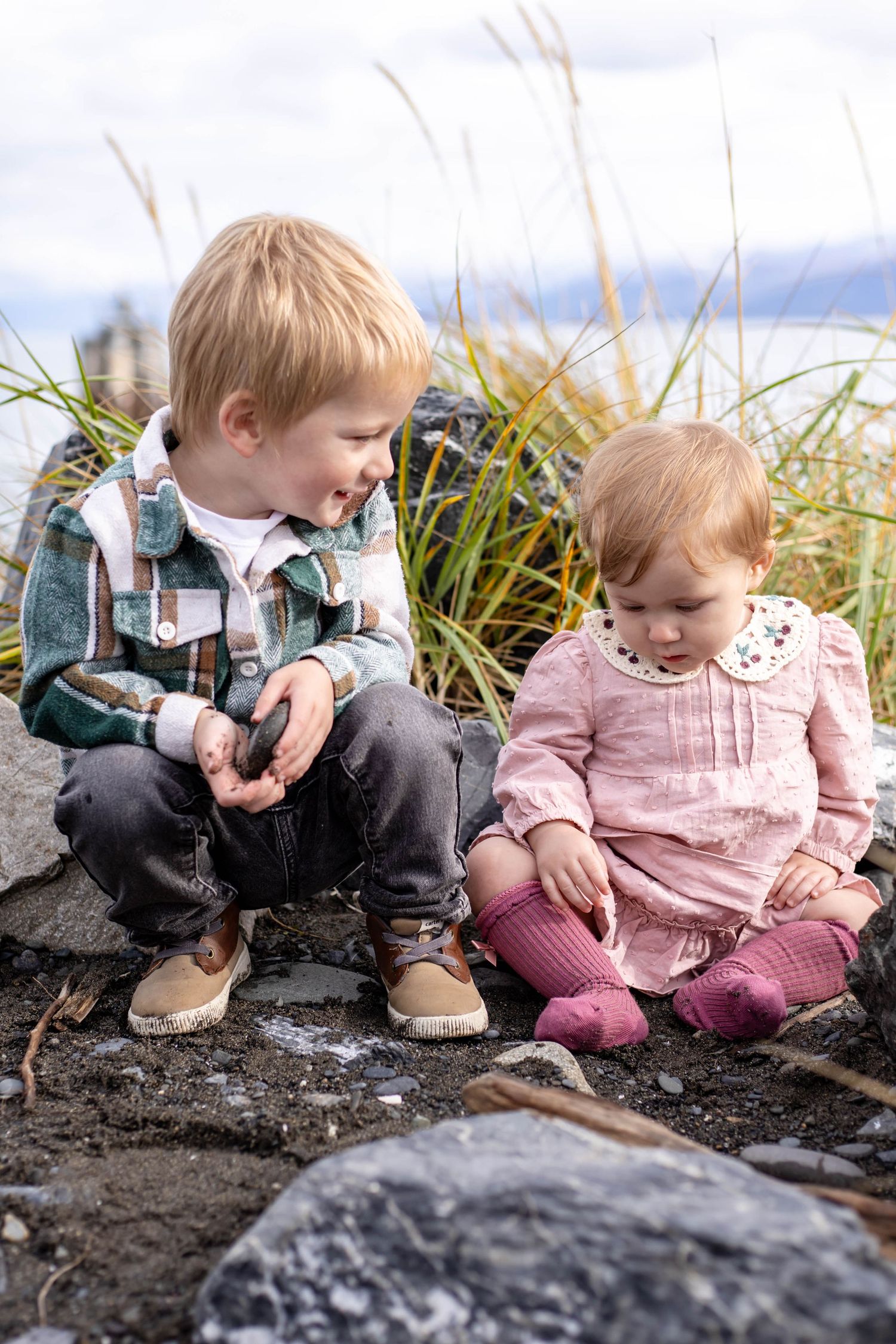 Two small children play together in muddy soil near rocks and beach grass.