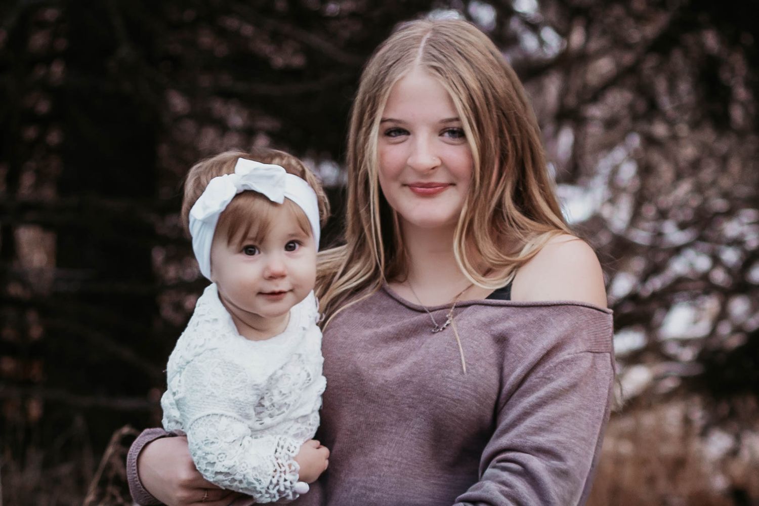 Teenage girl holding a baby dressed in white lace with a bow headband, posing outdoors.