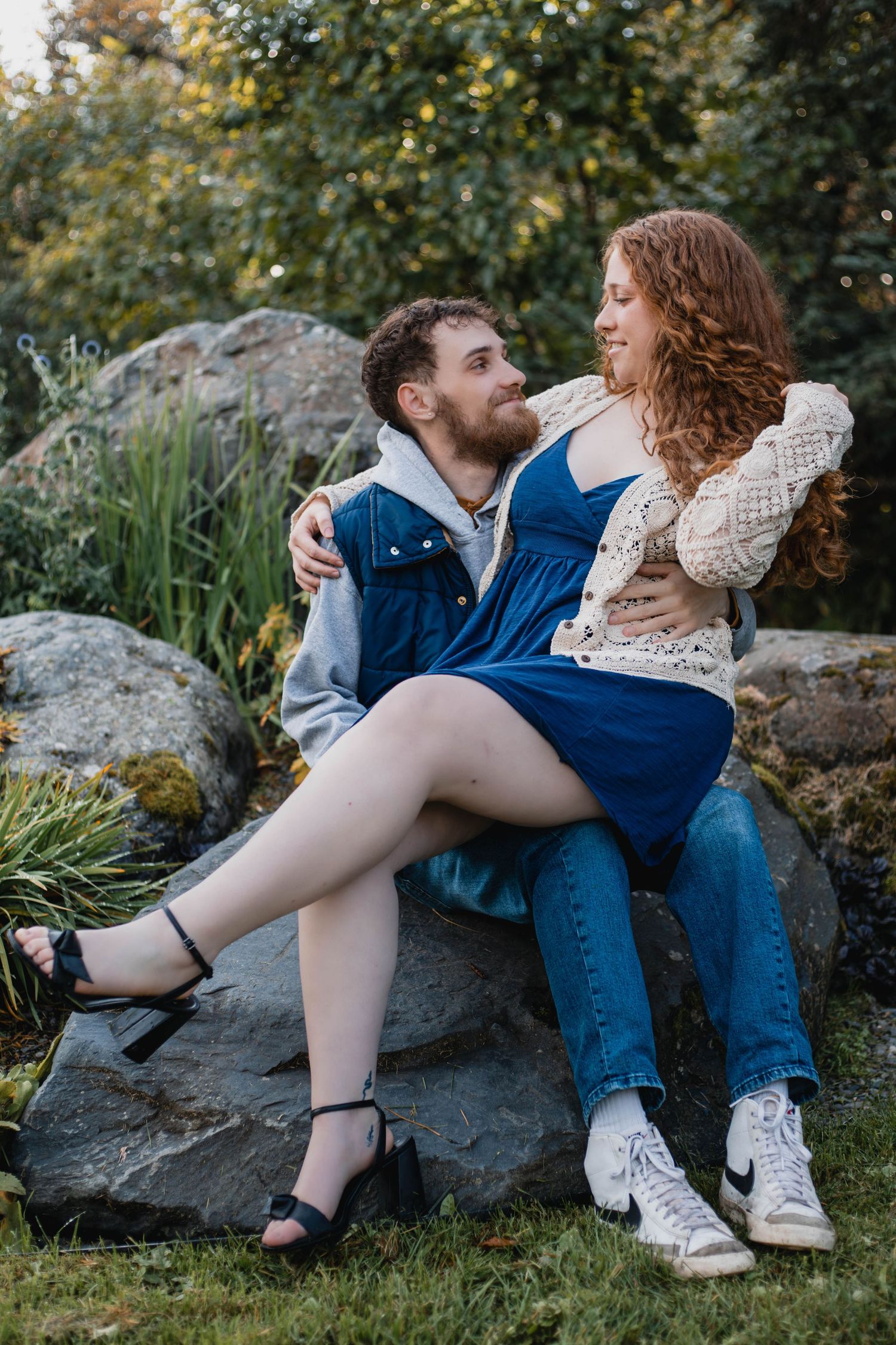 A couple sits together on a large rock in a garden setting wearing casual outfits.