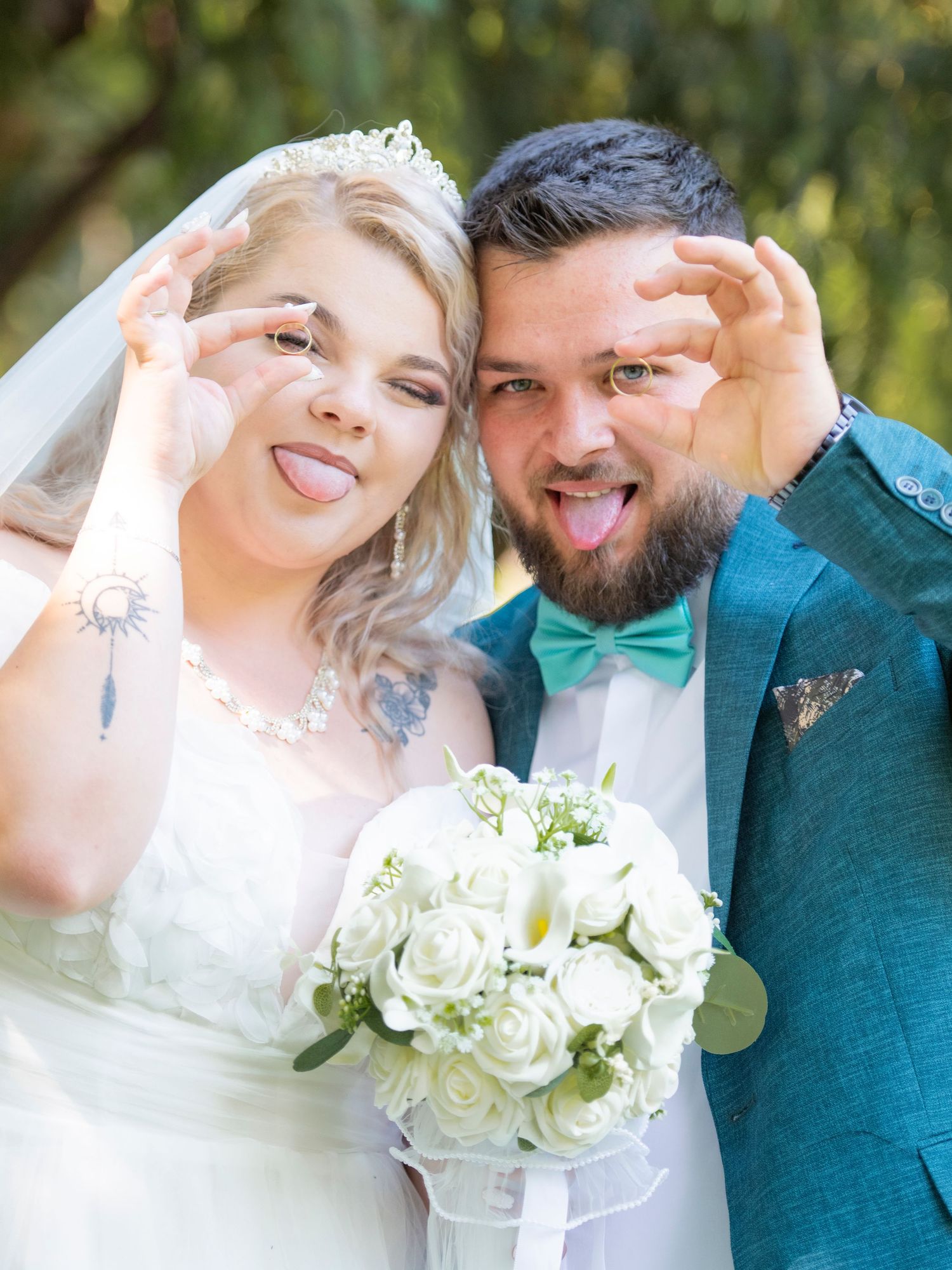 Newlywed couple in wedding attire playfully sticking tongues out, holding up rings, bride holding white rose bouquet.