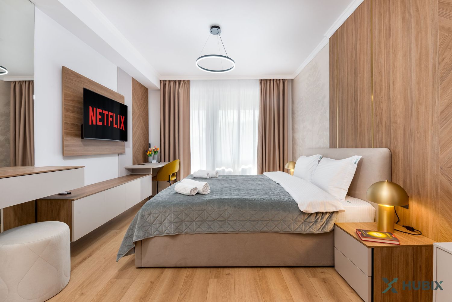 Modern hotel bedroom with Netflix TV, wooden accents, gray bedding, and golden lamp on hardwood floors.