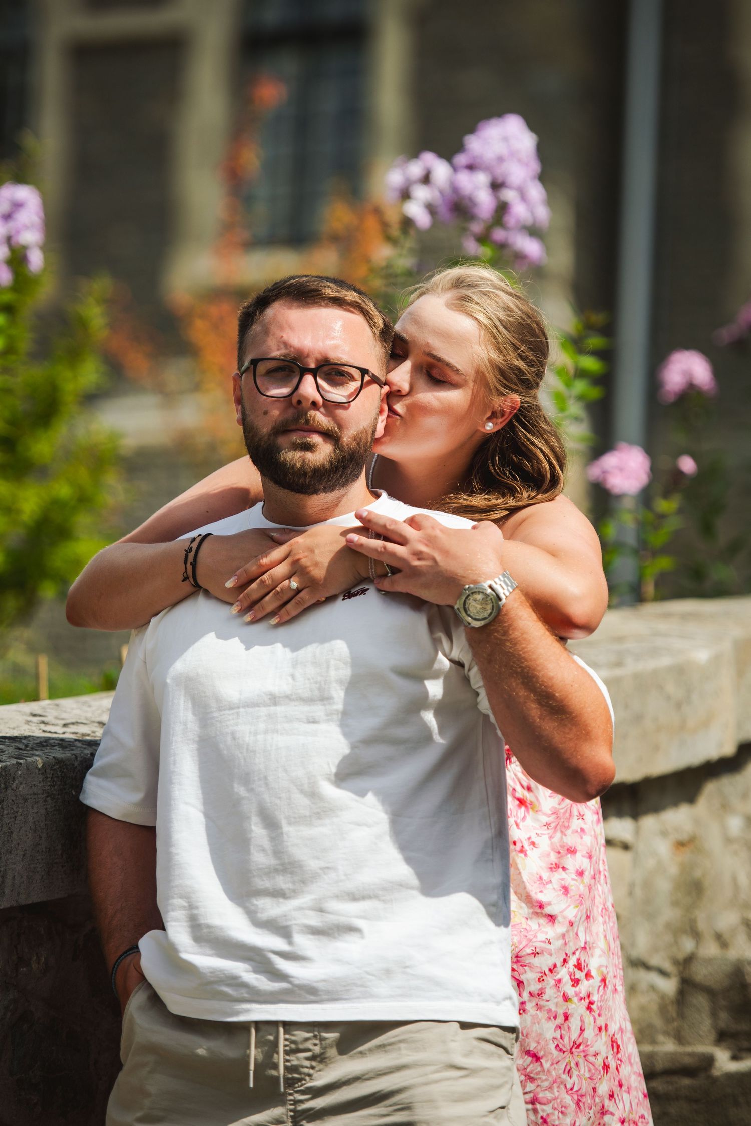 A woman in a floral dress kisses a bearded man wearing glasses outdoors near purple flowers.