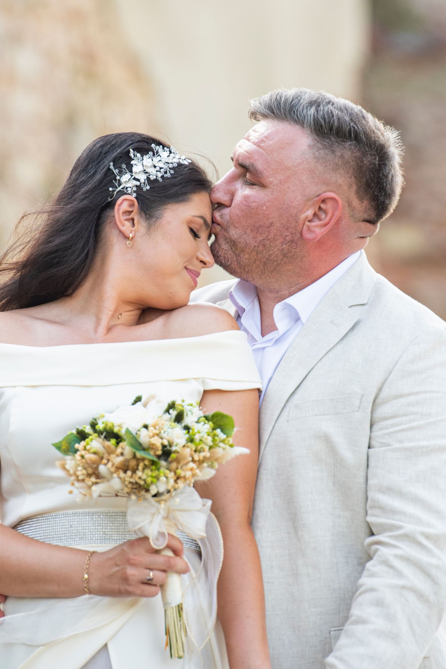 Groom in beige suit kissing bride on forehead, bride in white off-shoulder gown holding floral bouquet.