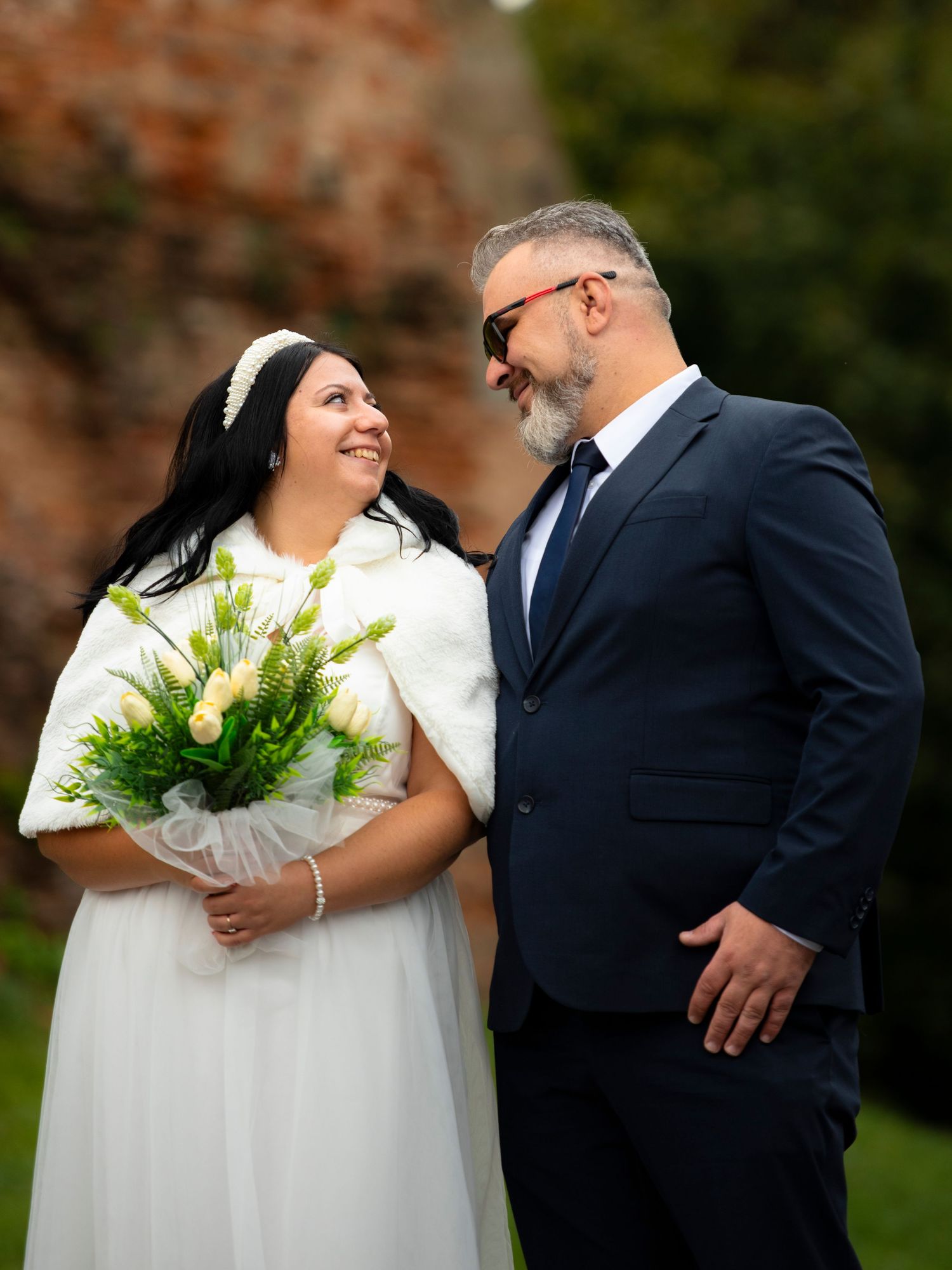 Bride in white dress holding green bouquet smiles at groom in navy suit outdoors at wedding ceremony.