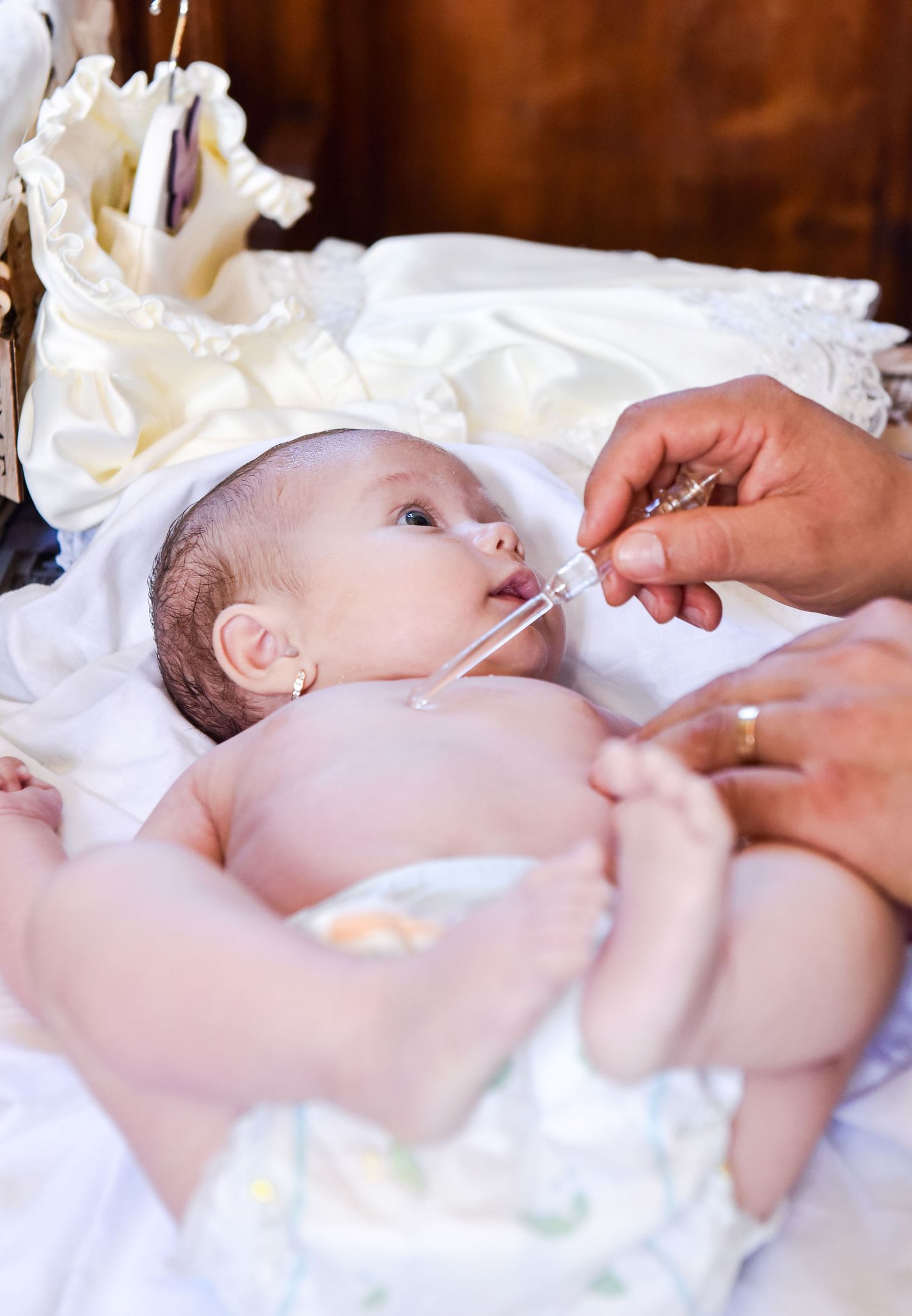 Newborn baby receiving liquid medicine via dropper during baptism ceremony, dressed in white christening gown.