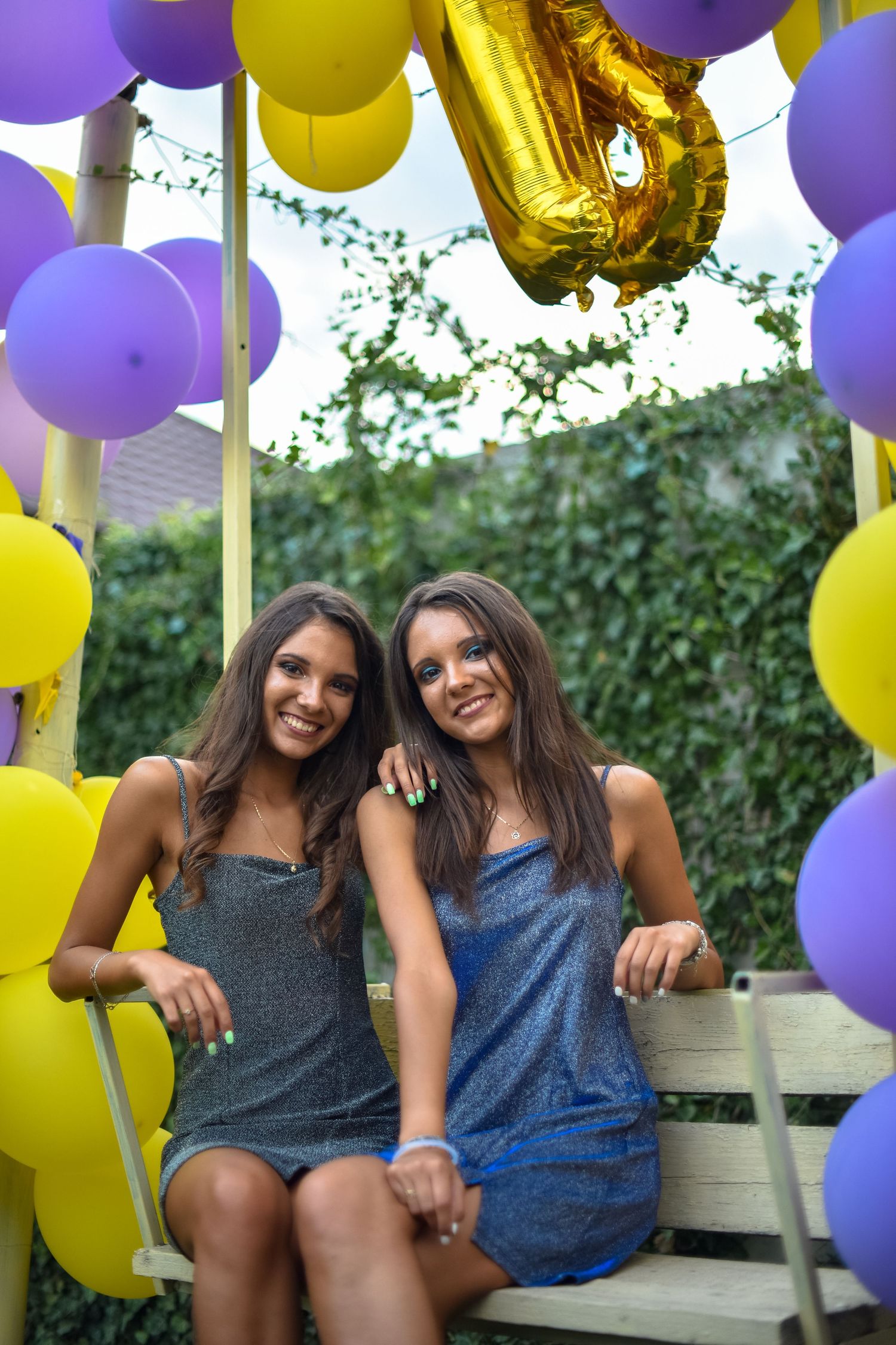 Two smiling young women in sparkly dresses sitting on a swing surrounded by purple and yellow balloons.