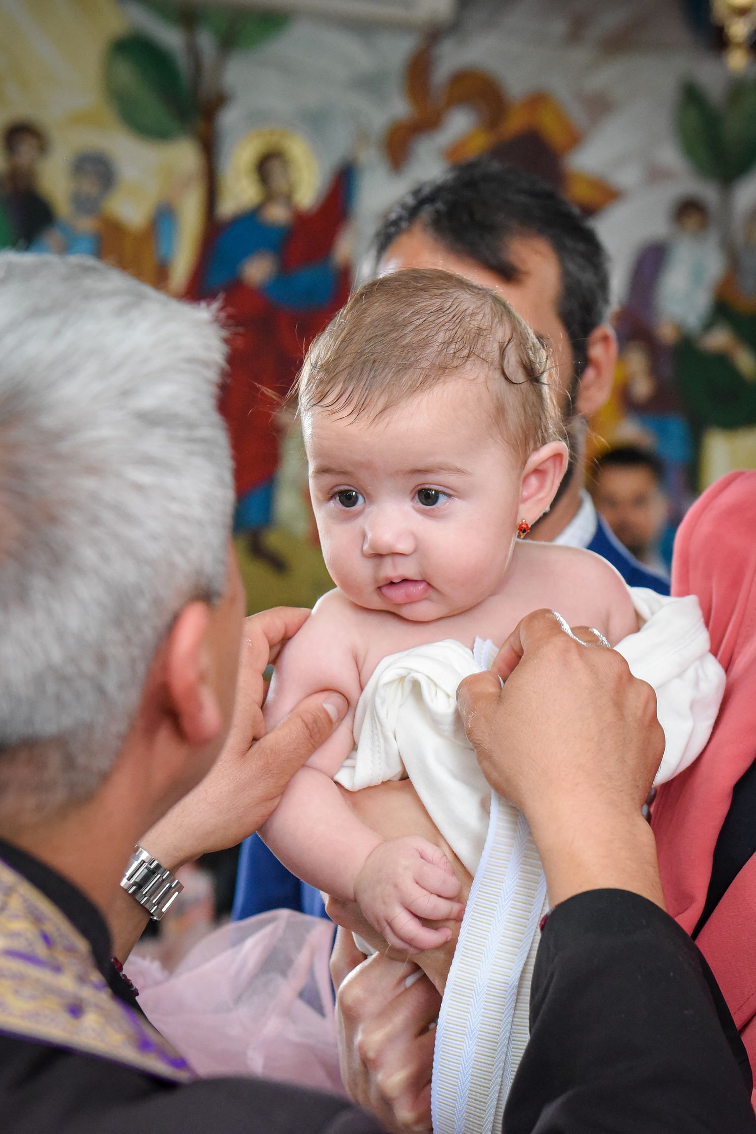 Priest performing Orthodox Christian baby baptism ceremony in church with religious icons in background.