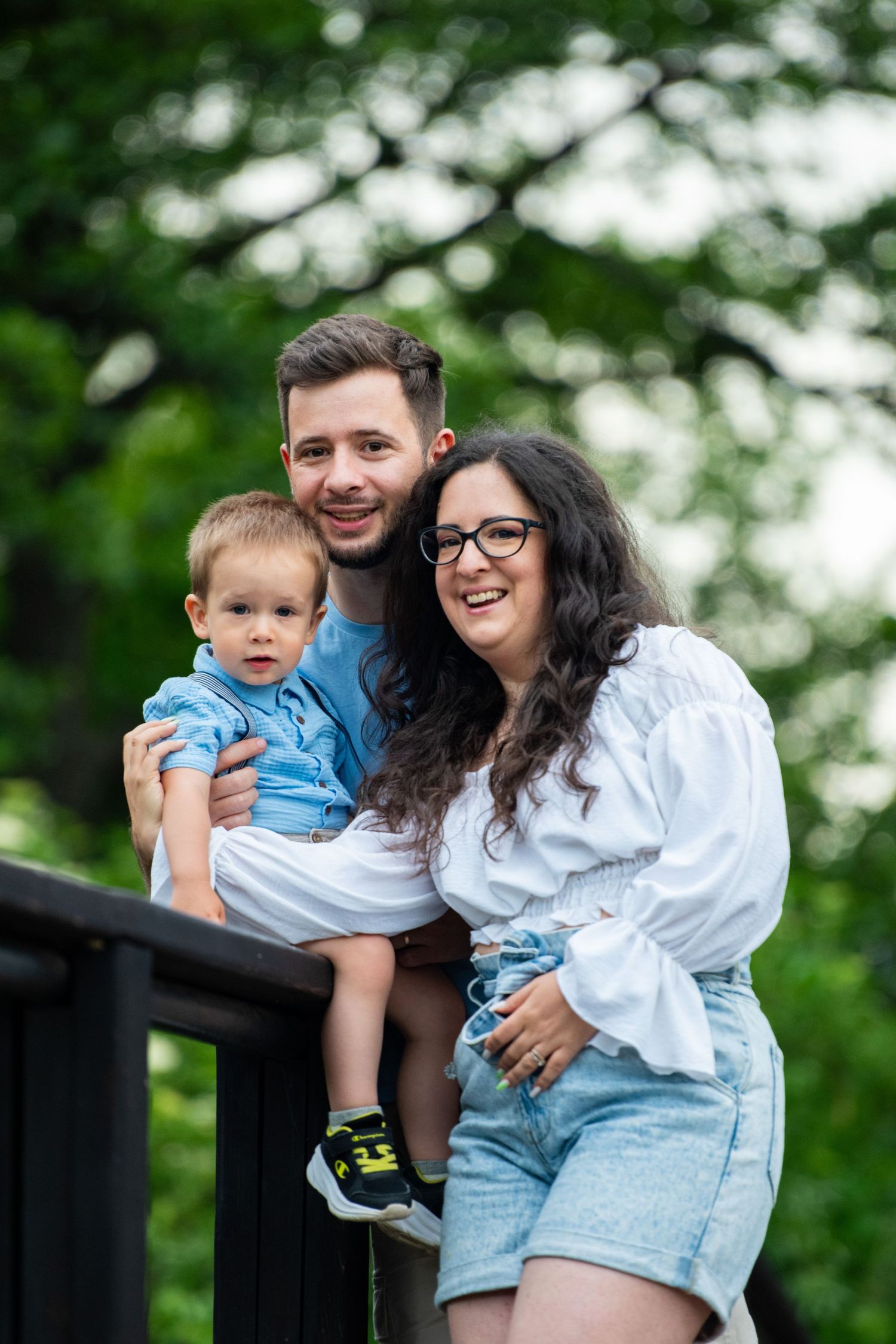 Happy young family with toddler boy posing on wooden deck surrounded by lush green trees outdoors.