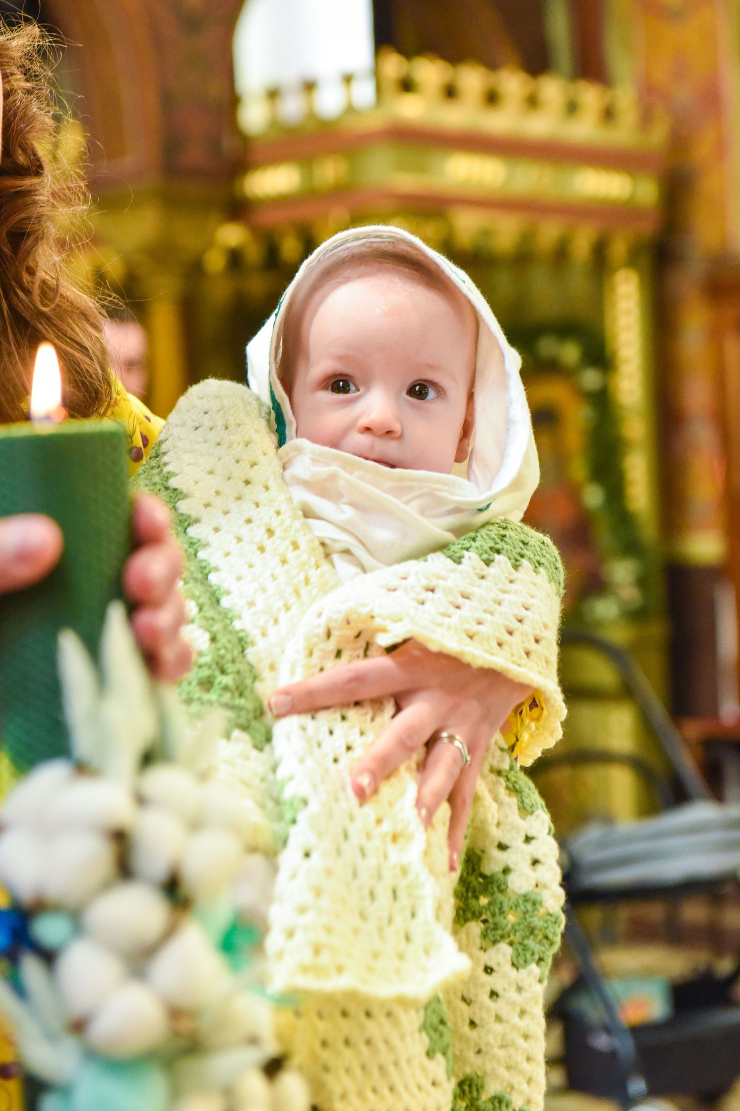 Baby wrapped in knitted yellow blanket with white headscarf held during Orthodox church baptism ceremony.