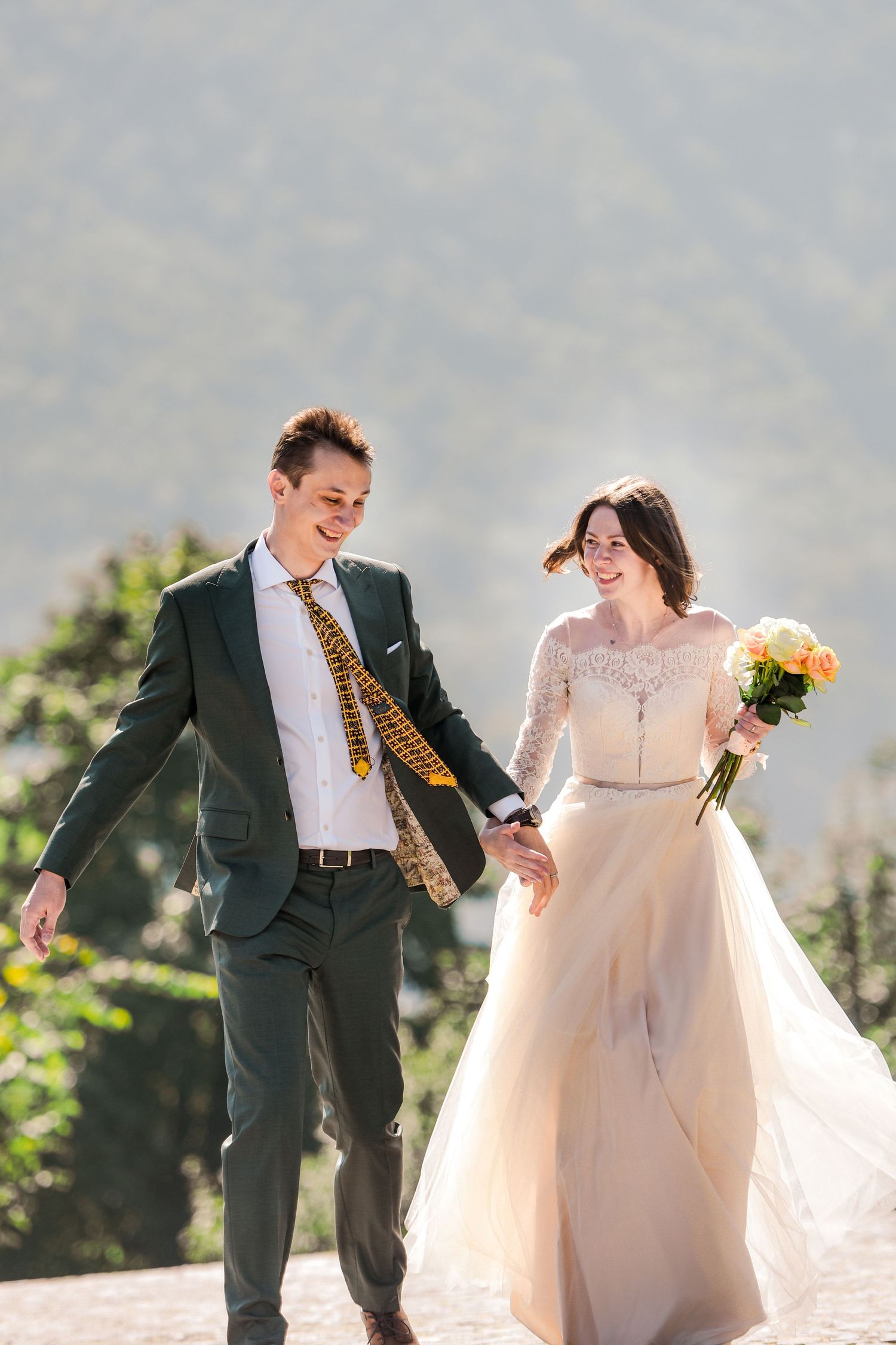 Happy newlyweds walk hand in hand outdoors, bride in champagne gown with bouquet, groom in dark suit with yellow tie.