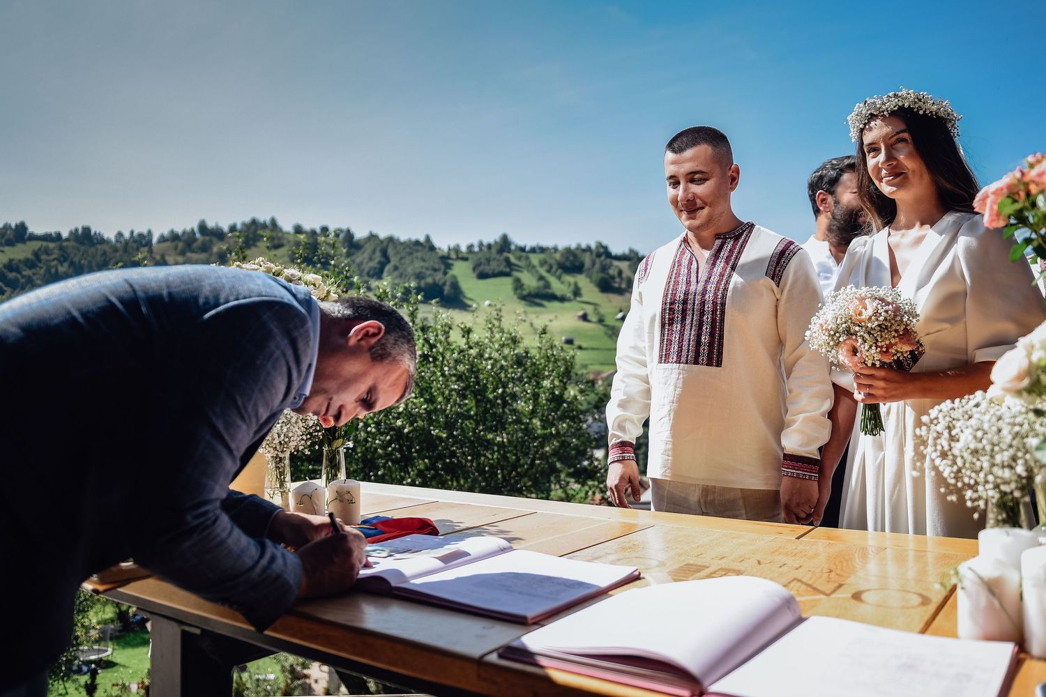 Official signing wedding register outdoors with couple in traditional attire and scenic green hills backdrop.