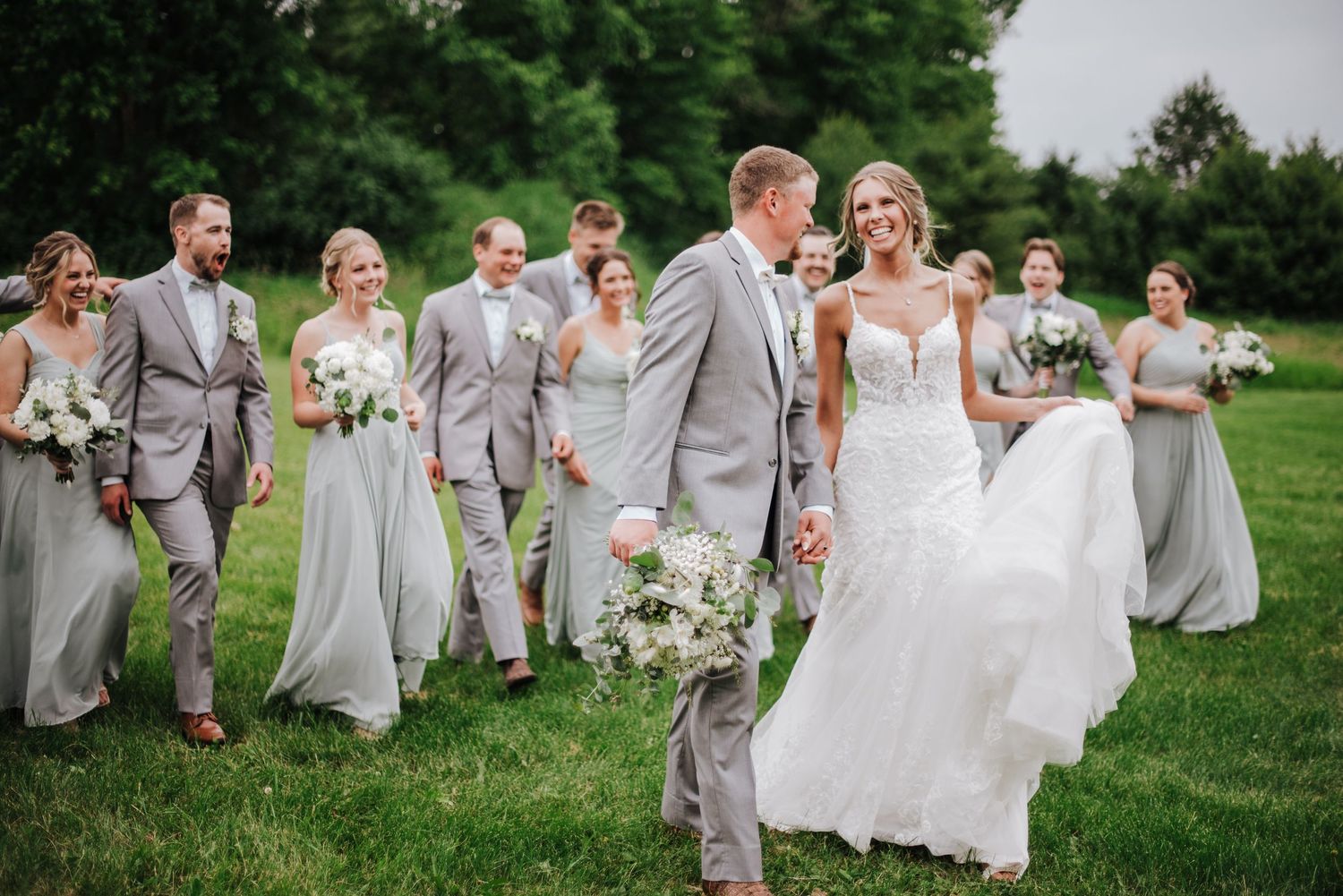 Wedding party in matching gray suits and sage bridesmaid dresses walking across green lawn together.